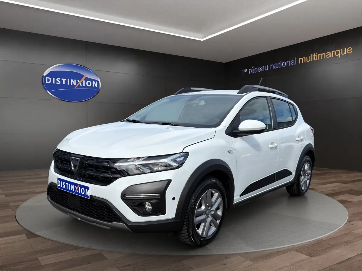 Dacia Sandero Stepway blanc vue 3/4 avant droit, avec barres de toit et protections noires sur les bas de caisse.