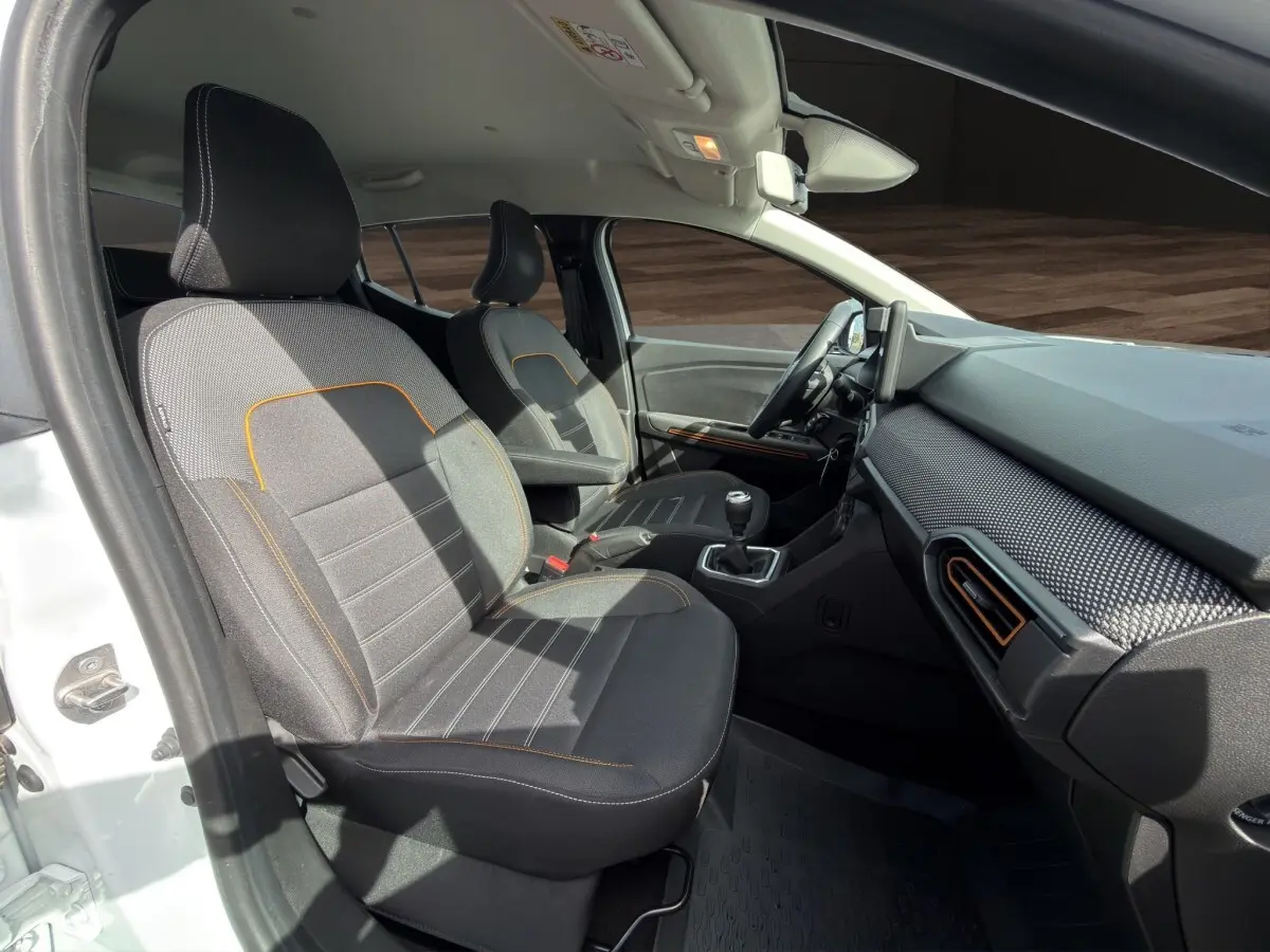 Vue intérieure côté conducteur de la Dacia Sandero Stepway blanche, sièges gris avec surpiqûres orange et tableau de bord moderne.