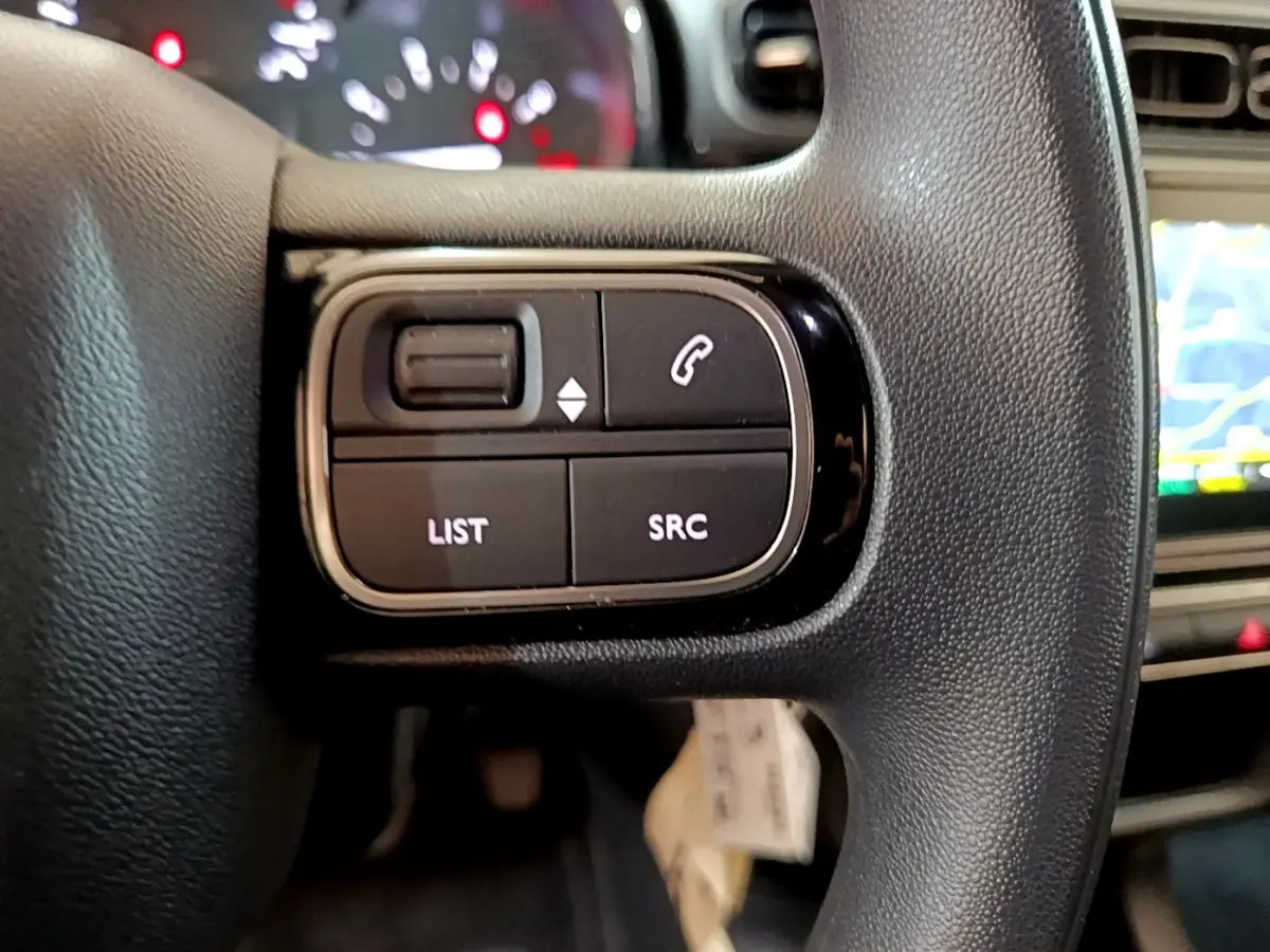 Gros plan sur les commandes gauche du volant noir de la Citroën C3 blanche, avec tableau de bord et écran tactile en arrière-plan.