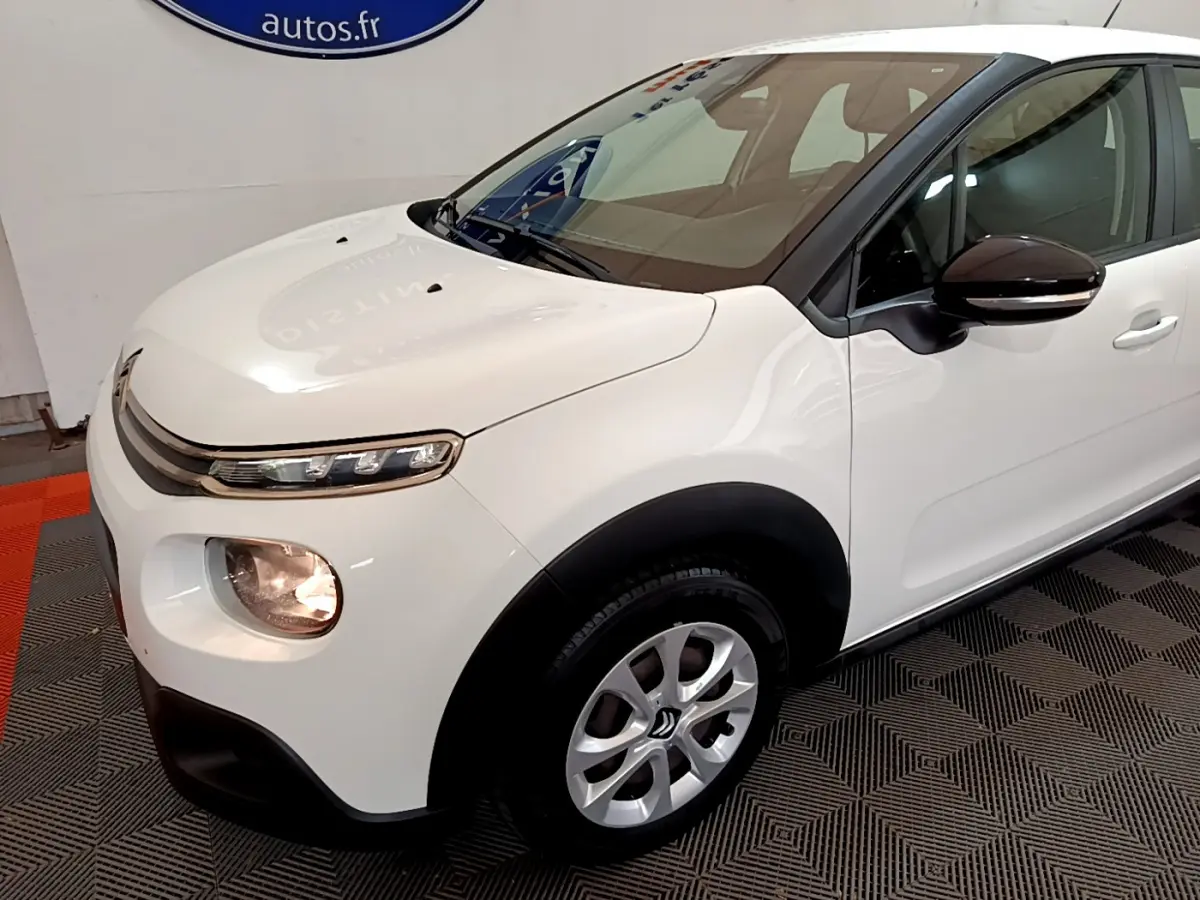 Vue 3/4 avant droit d'une Citroën C3 blanche avec rétroviseurs noirs et jantes alliage en intérieur showroom.