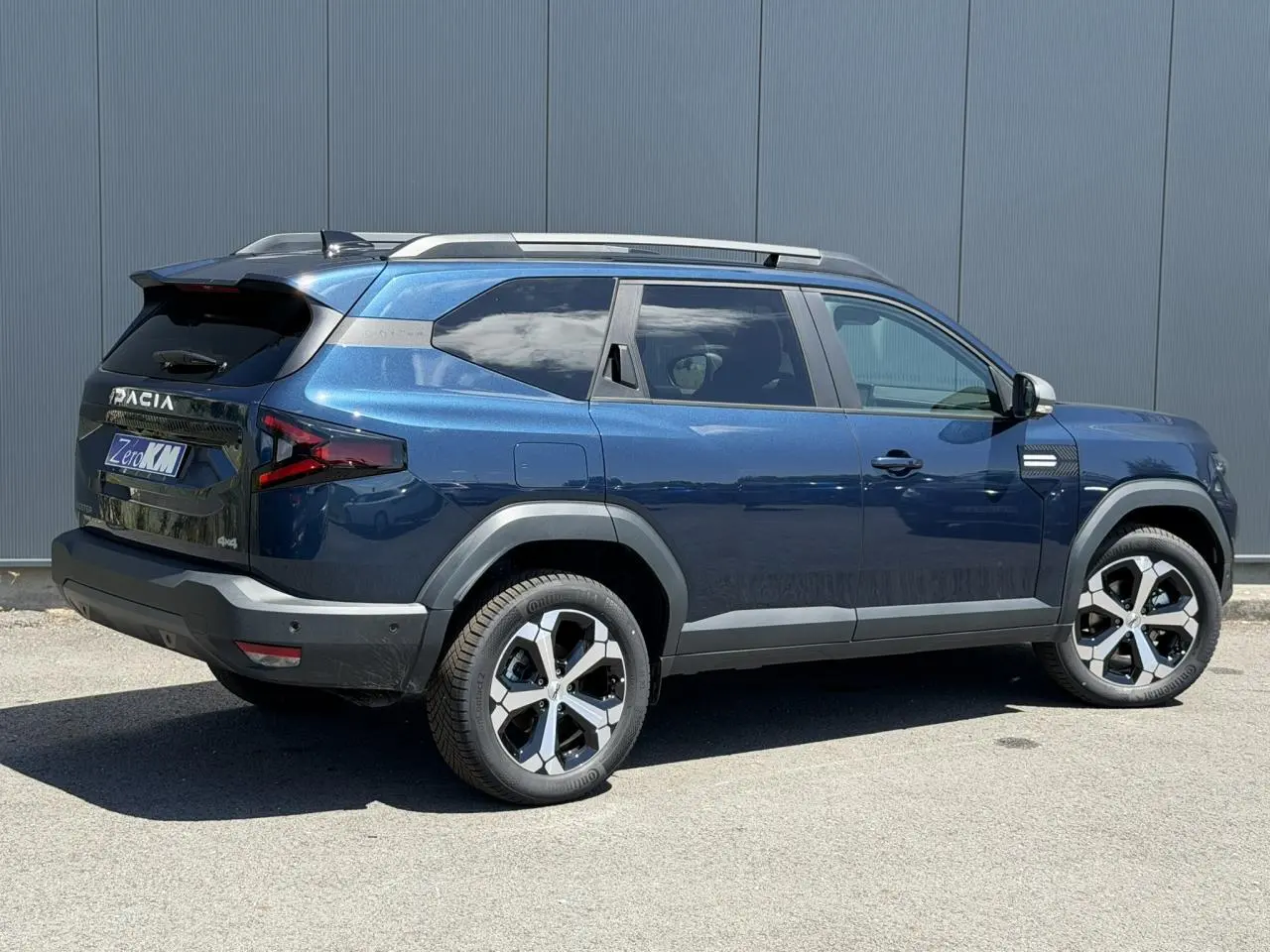 Vue 3/4 arrière droite du SUV Dacia Bigster bleu indigo avec jantes alliage 18 pouces et barres de toit grises.