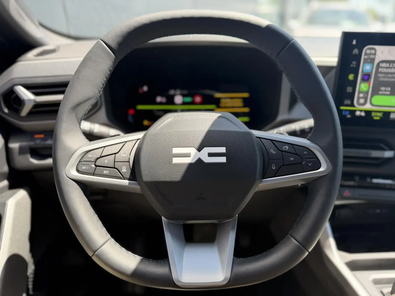 Vue rapprochée du volant noir du Dacia Bigster Hybrid-G 150 4X4 avec commandes intégrées et tableau de bord numérique flou.