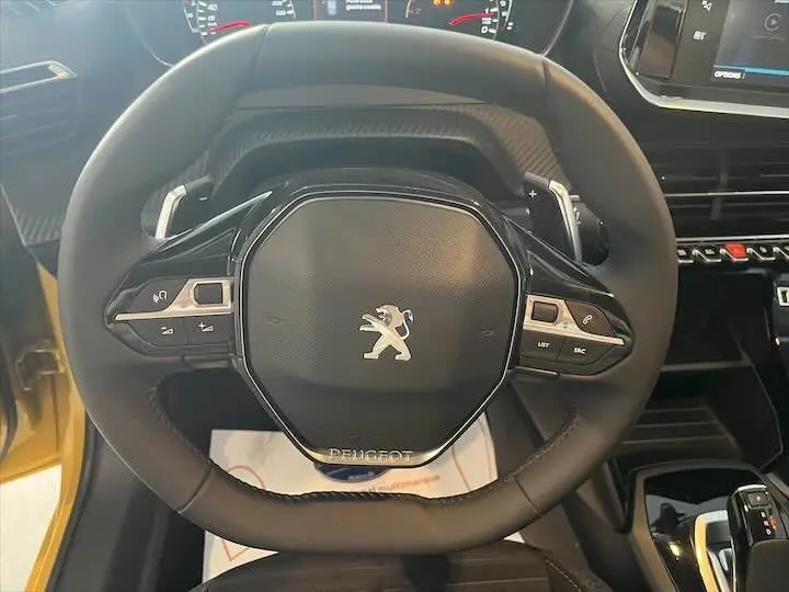 Vue rapprochée du volant noir du Peugeot 208 jaune Faro Metal, avec commandes intégrées et tableau de bord partiellement visible.