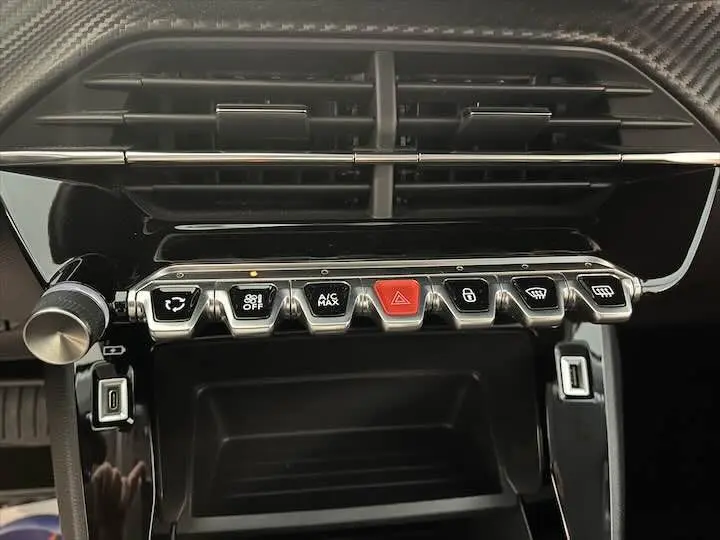 Gros plan sur la console centrale de la Peugeot 208 2021, montrant les boutons de commande sous la grille d'aération noire.
