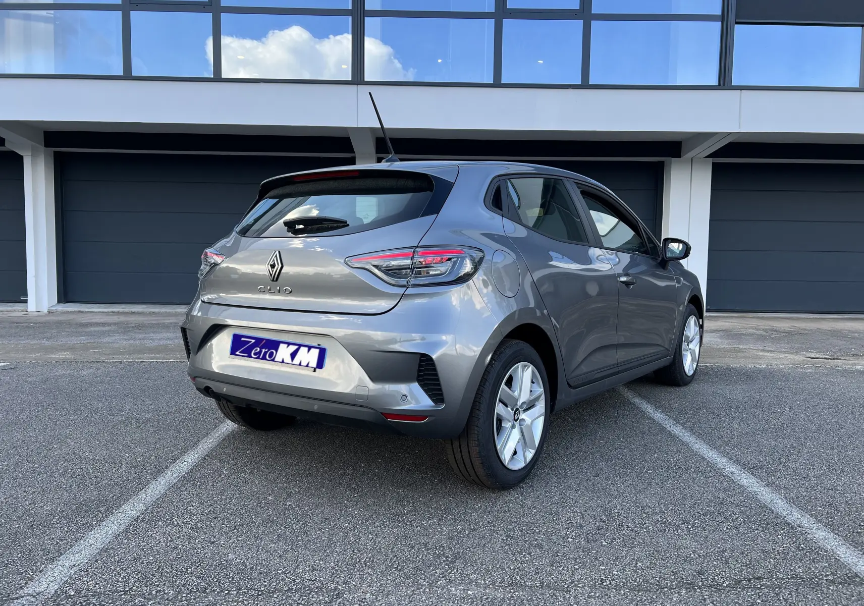 Renault Clio V gris schiste vue 3/4 arrière droit, jantes alliage 16 pouces et protections latérales chromées visibles.