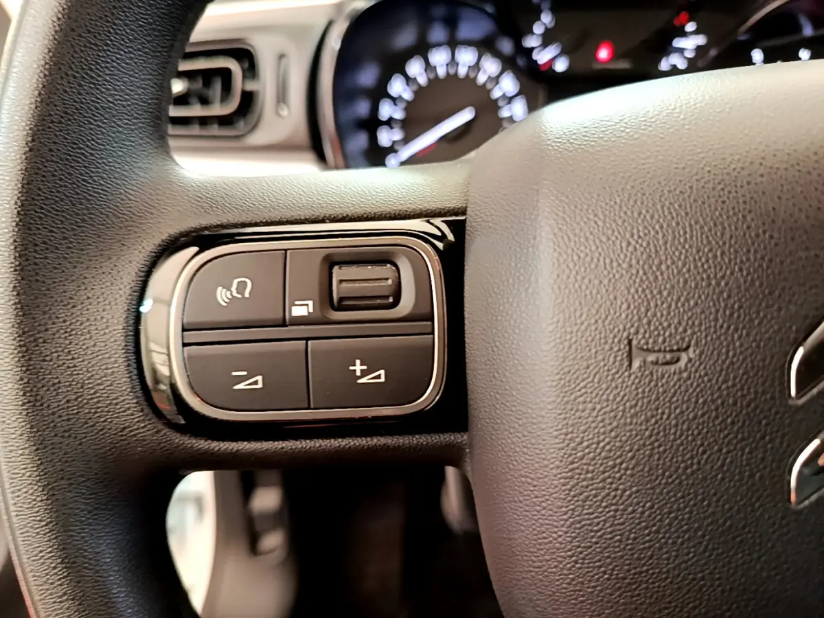 Gros plan sur les commandes gauche du volant noir d'une Citroën C3 blanche, tableau de bord flou en arrière-plan.