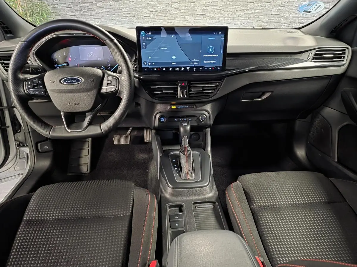 Vue intérieure avant de la Ford Focus 2024 ST Line, tableau de bord noir avec écran tactile central et volant sport à surpiqûres rouges.