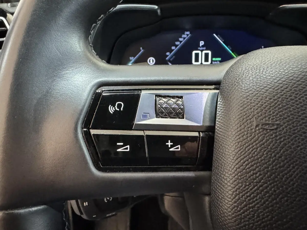 Gros plan sur les commandes gauche du volant cuir du DS7 Crossback gris foncé, avec affichage numérique en arrière-plan.