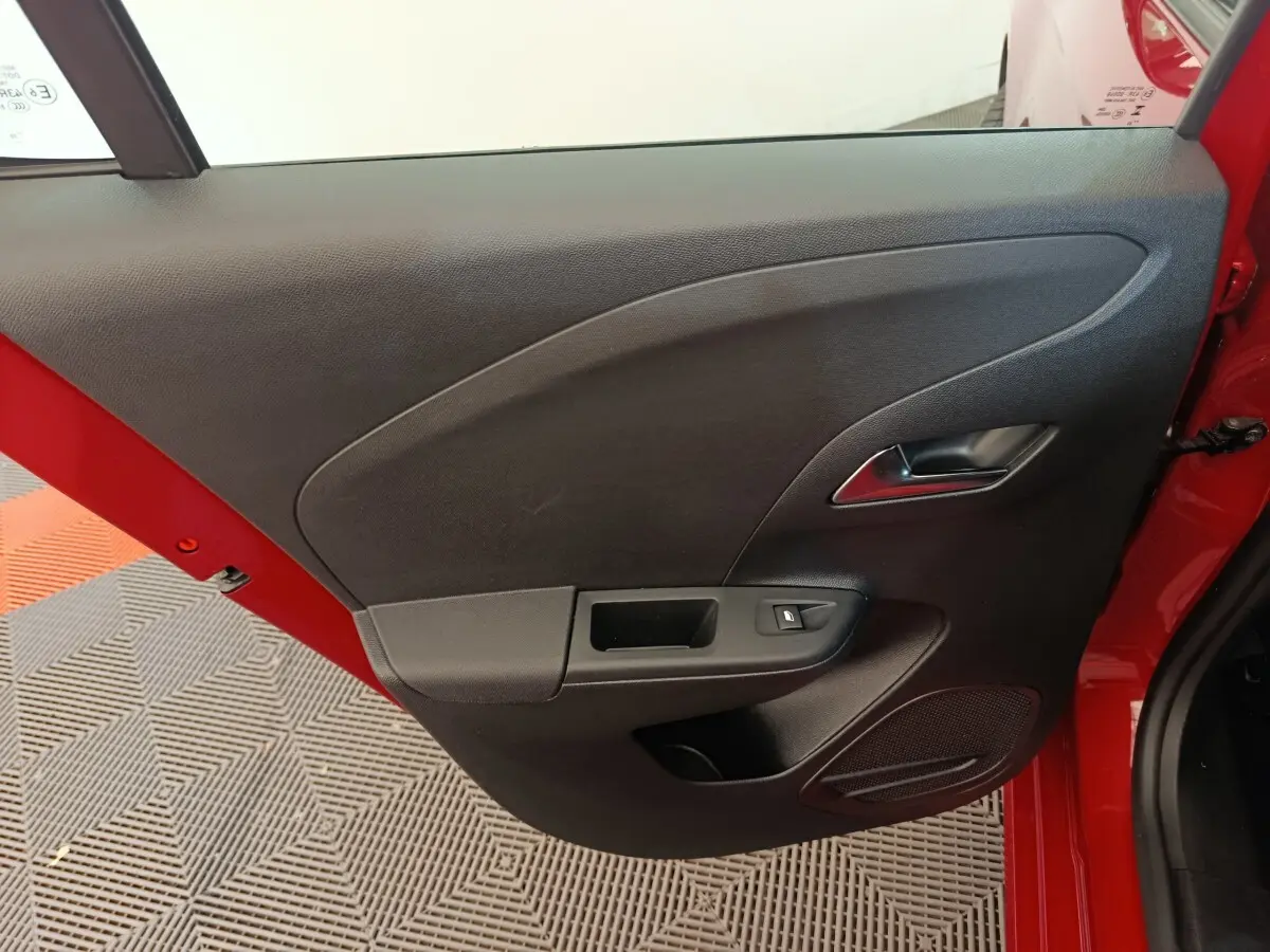 Vue rapprochée de la porte intérieure côté gauche d’une Opel Corsa rouge, avec poignée argentée et commande de fenêtre.