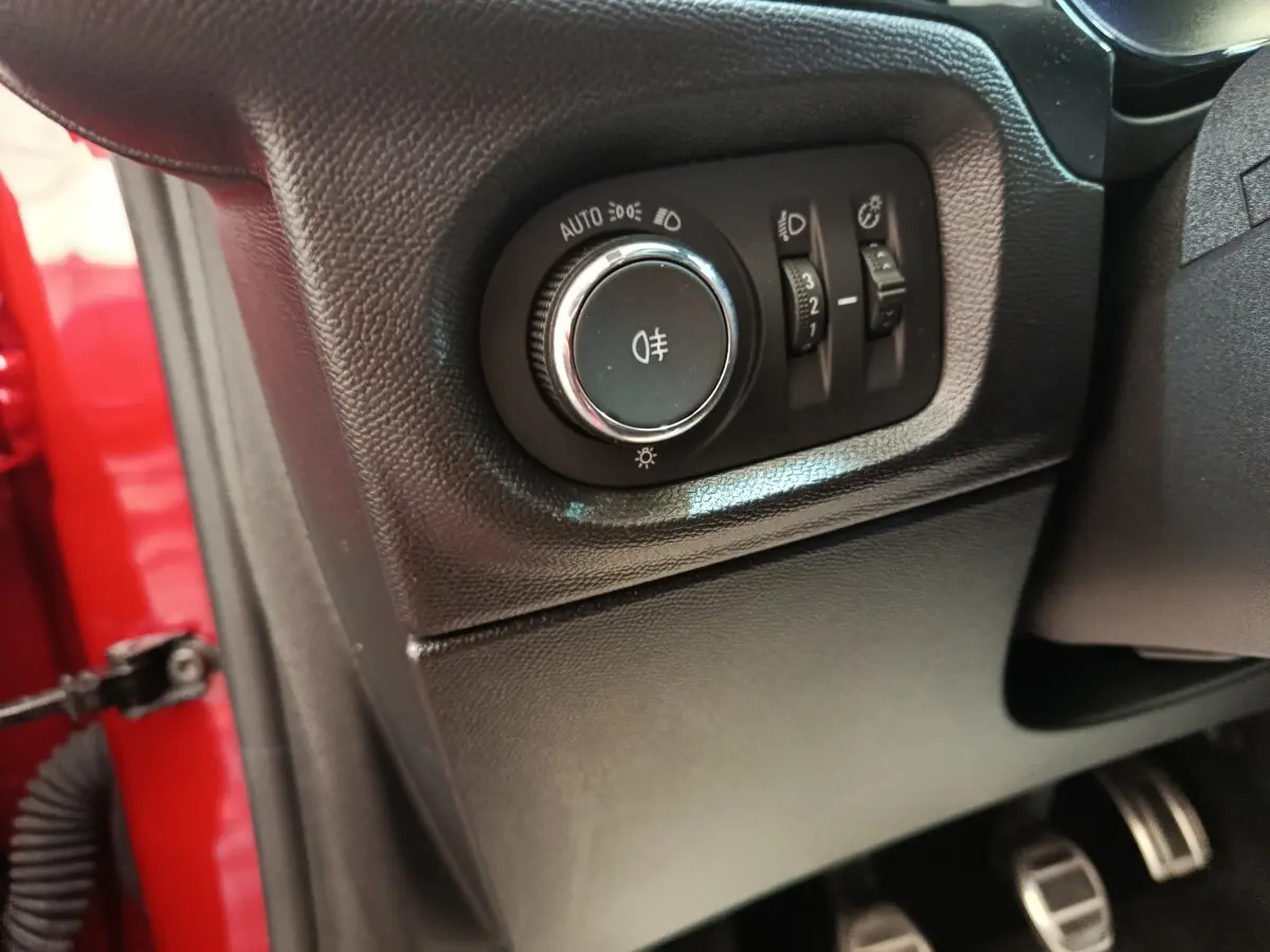 Gros plan sur le panneau de commande des phares à gauche du volant, intérieur noir, portière rouge visible.