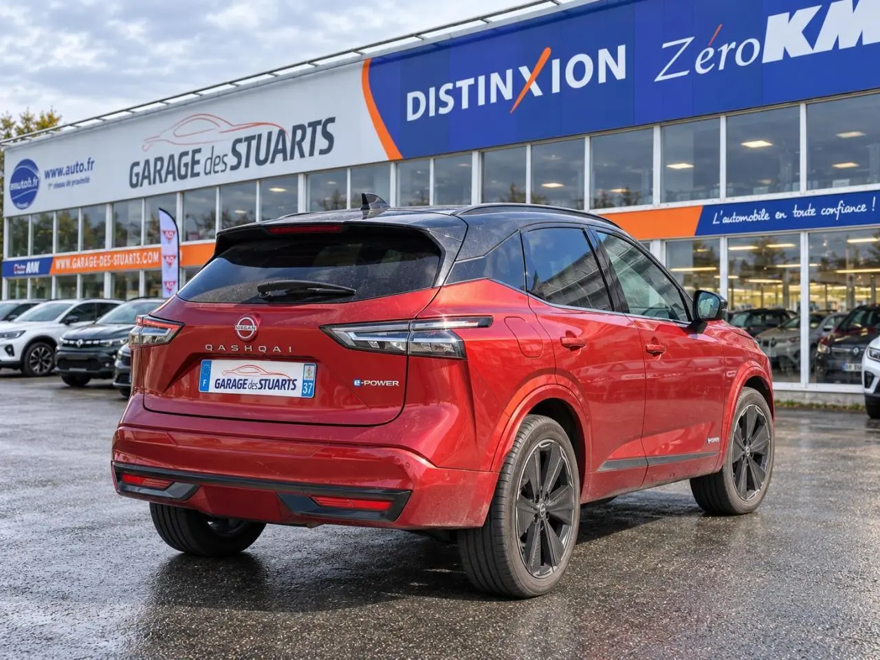 Vue 3/4 arrière d’un Nissan Qashqai 2024 rouge foncé avec toit noir et jantes noires, stationné devant un garage.