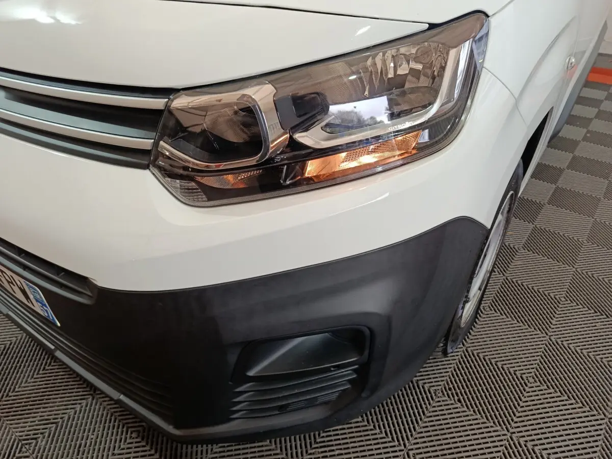 Gros plan sur le phare avant droit allumé du Citroën Berlingo blanc, avec pare-chocs noir et partie de la roue visible.