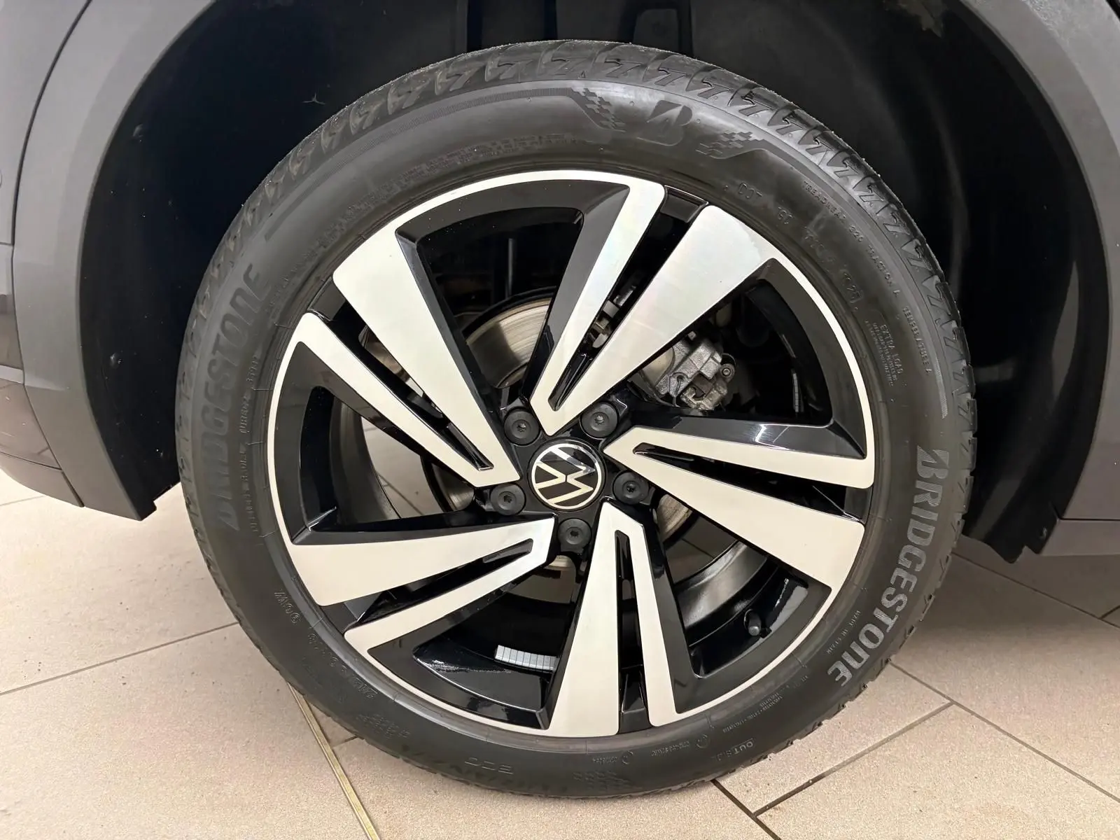 Gros plan sur la roue avant droite du Volkswagen T-Roc noir intense, mettant en valeur la jante alliage bicolore et le pneu Bridgestone.