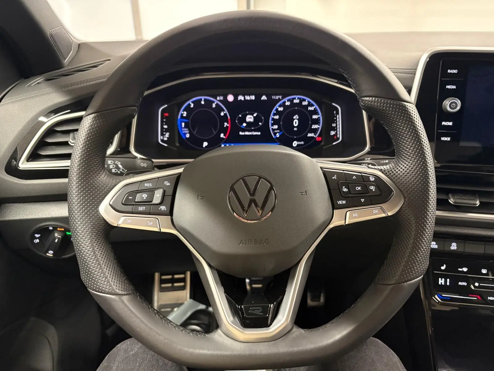 Vue rapprochée du volant noir perforé du Volkswagen T-Roc 1.5 TSI R-Line avec tableau de bord numérique allumé.