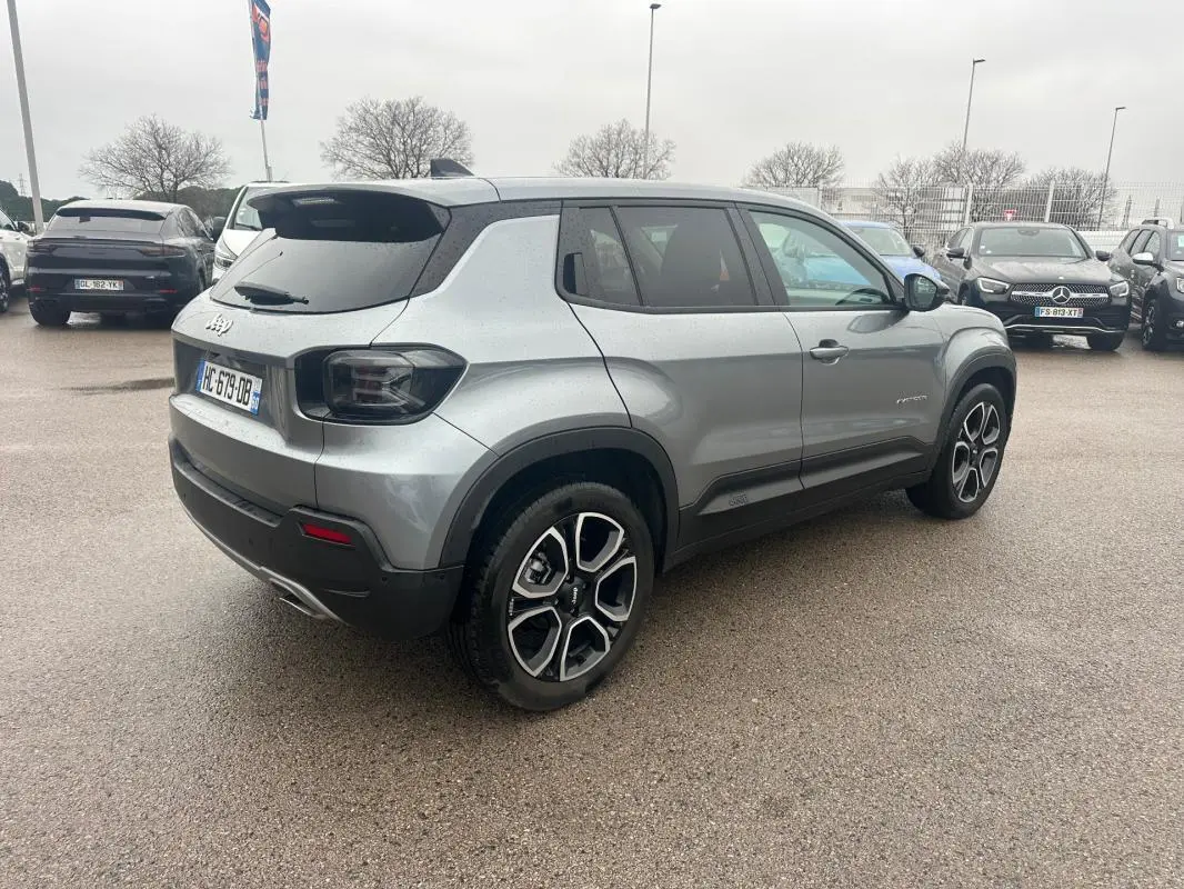Vue 3/4 arrière droite d’un Jeep Avenger gris foncé avec jantes alliage 18 pouces et toit noir sur parking.