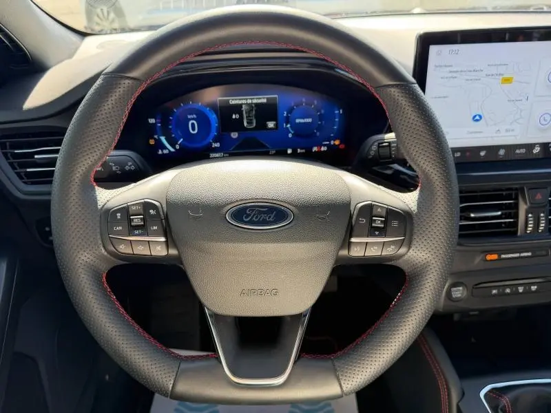 Vue intérieure centrée sur le volant perforé avec surpiqûres rouges de la Ford Focus SW Bleu Azur 2023, tableau de bord numérique et écran tactile visibles.
