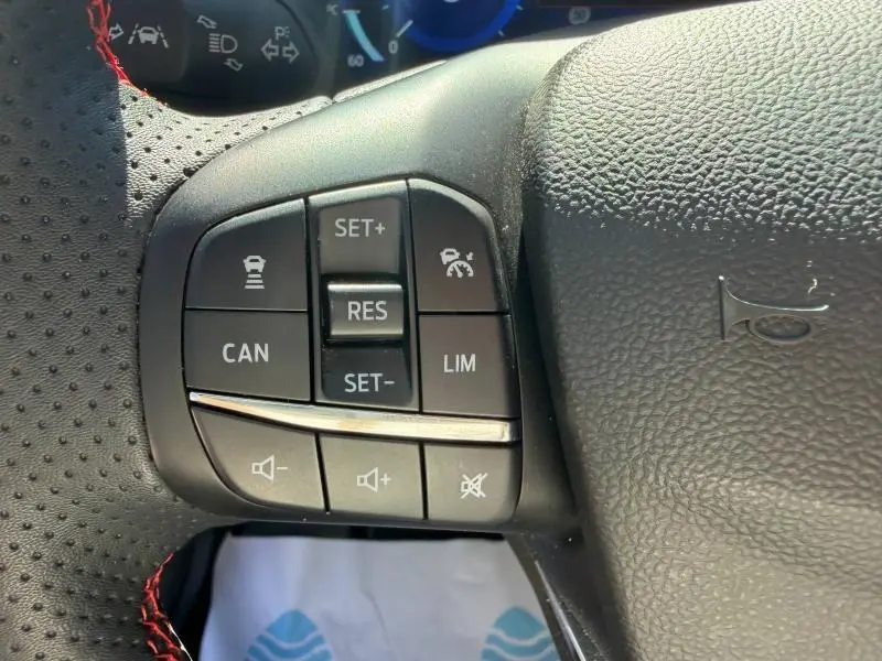 Gros plan sur les commandes de régulateur et volume au volant d'une Ford Focus SW 2023, cuir perforé avec surpiqûres rouges.
