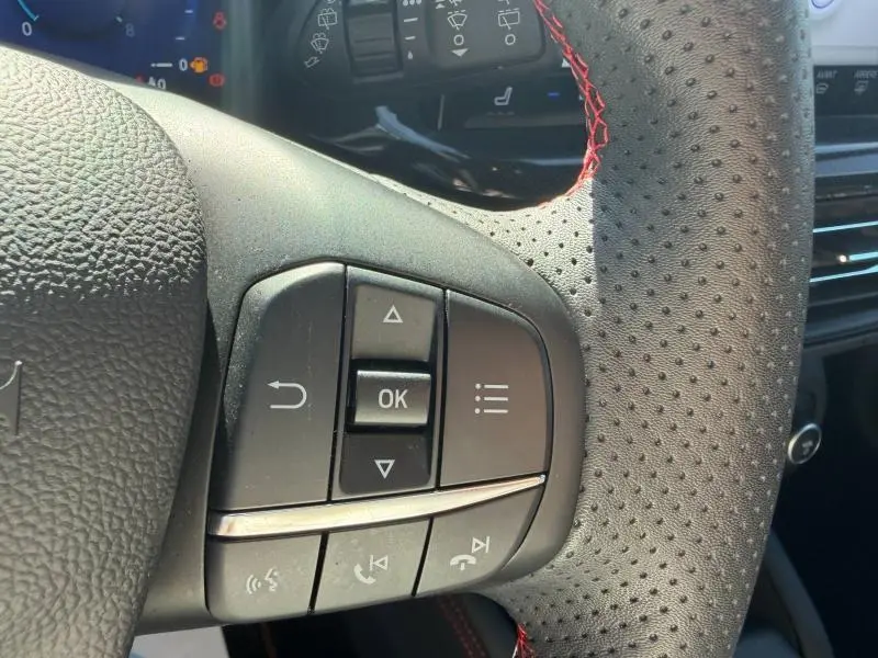 Gros plan sur les commandes multifonctions à gauche du volant perforé avec surpiqûres rouges d'une Ford Focus SW Bleu Azur 2023.