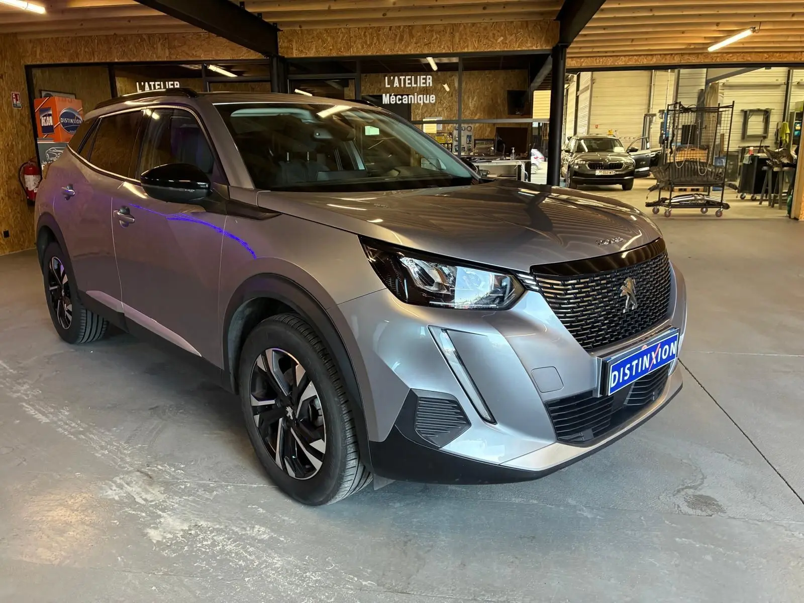 Peugeot 2008 gris en 3/4 avant droit dans un atelier mécanique, avec calandre noire et jantes alliage.