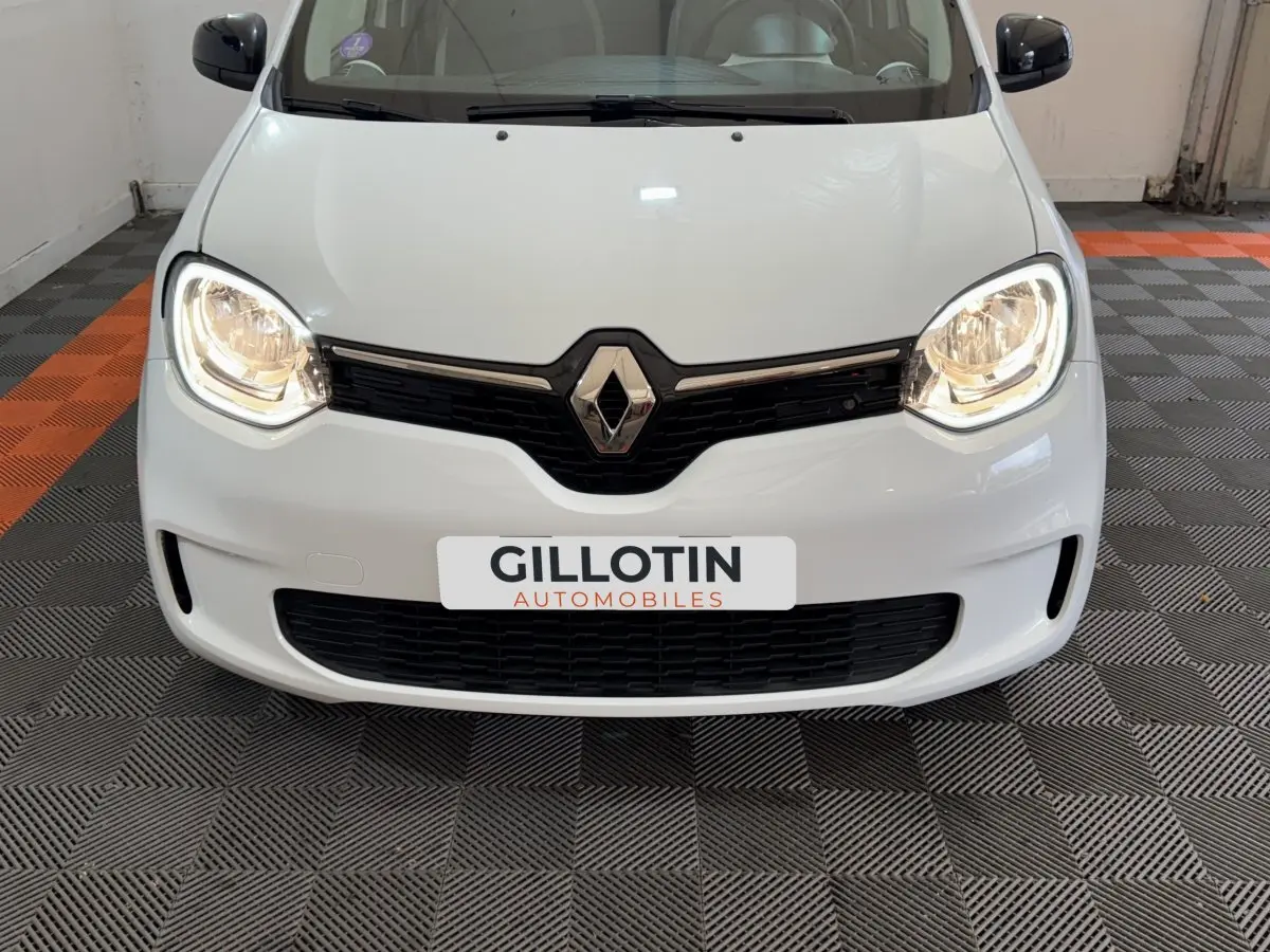 Vue frontale rapprochée de la Renault Twingo blanche avec phares allumés dans un garage.