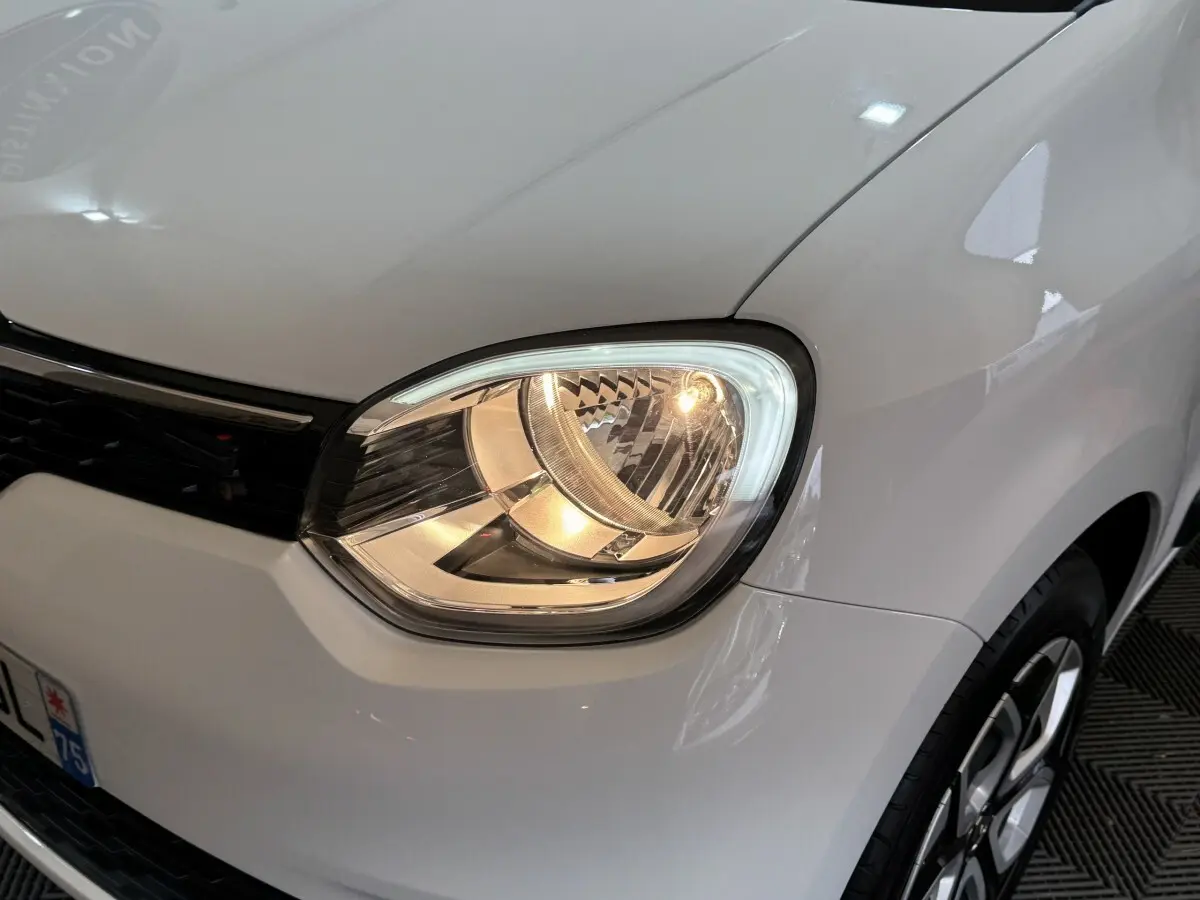 Gros plan sur le phare avant droit allumé d'une Renault Twingo blanche 2022, avec partie de la calandre visible.
