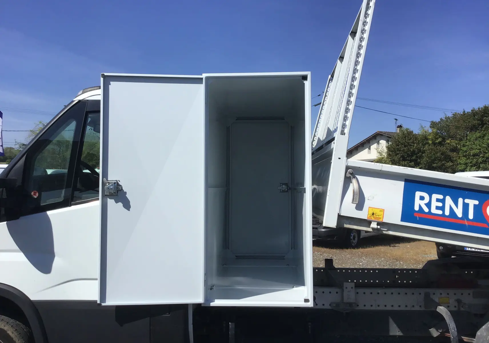 Vue latérale droite d’un Iveco Daily blanc avec benne basculante et coffre de rangement ouvert sur fond de ciel bleu.