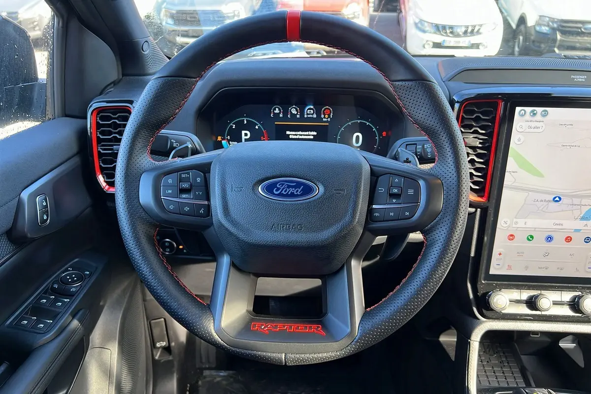 Vue intérieure centrée sur le volant cuir noir avec logo Ford et inscription rouge Raptor, tableau de bord digital et écran tactile.