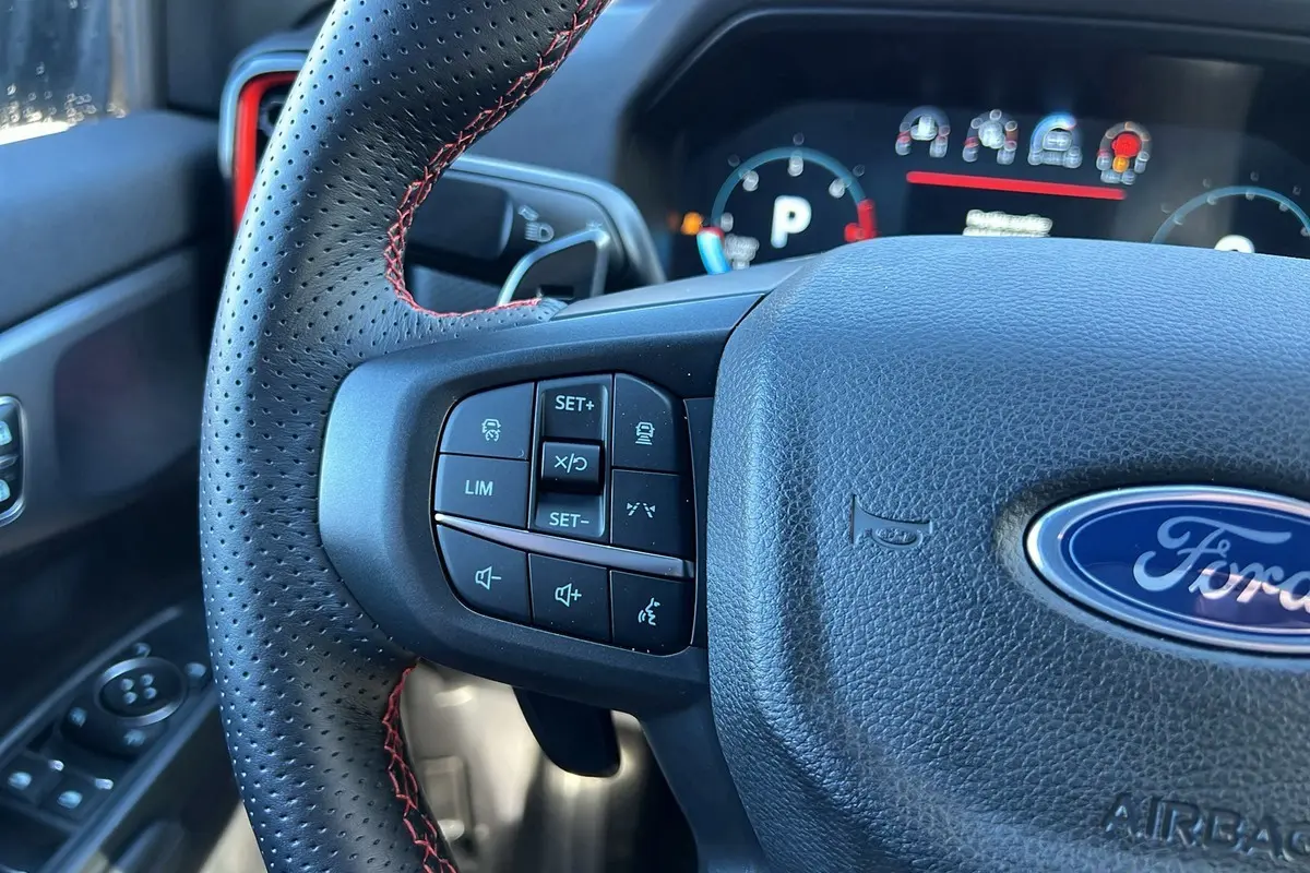 Gros plan sur le volant cuir perforé noir avec surpiqûres rouges du Ford Ranger Raptor 2025, boutons multifonctions visibles.