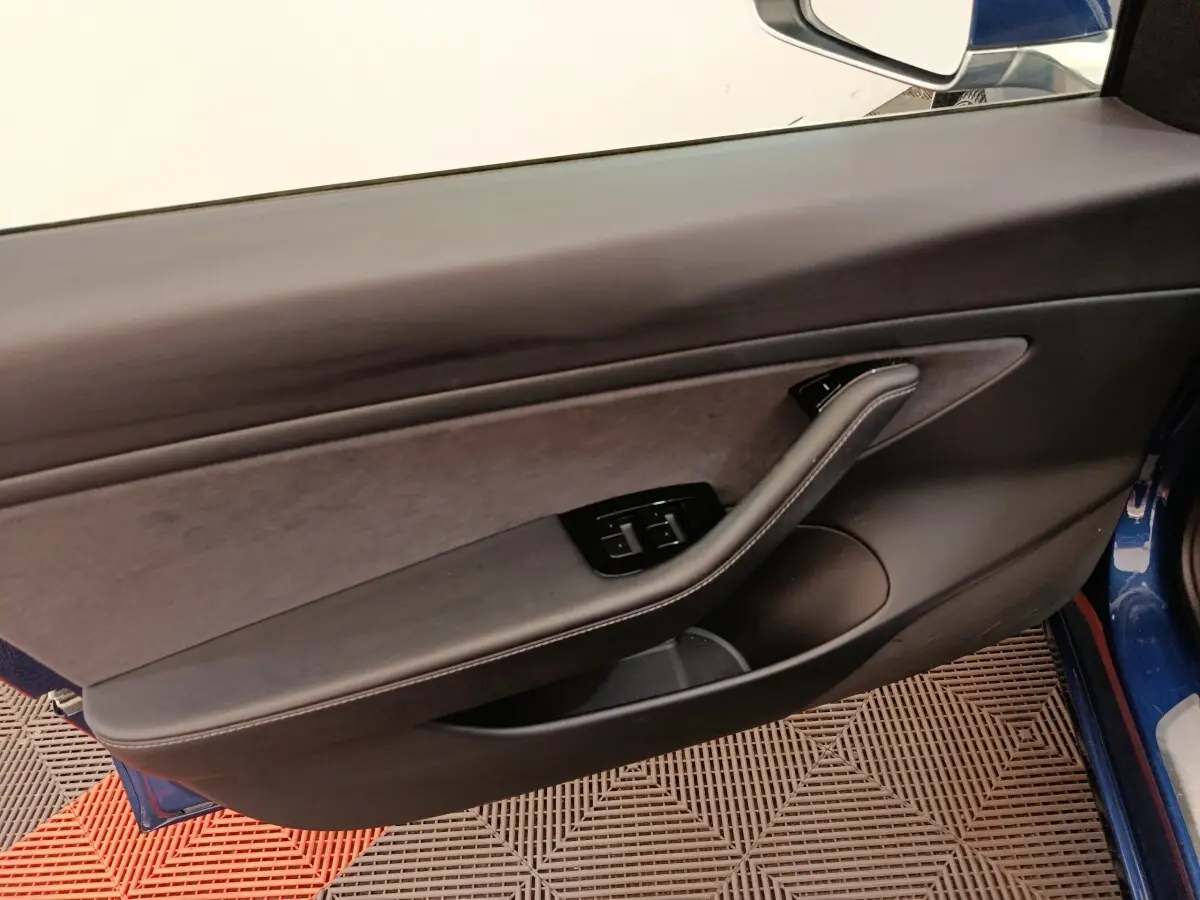 Vue intérieure sur la porte côté gauche d’une Tesla Model 3 bleu, avec commandes de vitres et poignée intégrée.