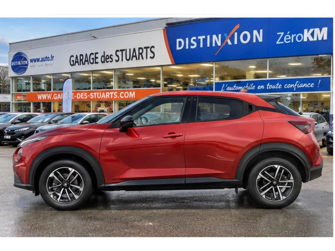 Vue latérale gauche d'un Nissan Juke 1.6 Hybrid rouge Fuji Sunset avec jantes alliage 17 pouces, garé devant un garage.