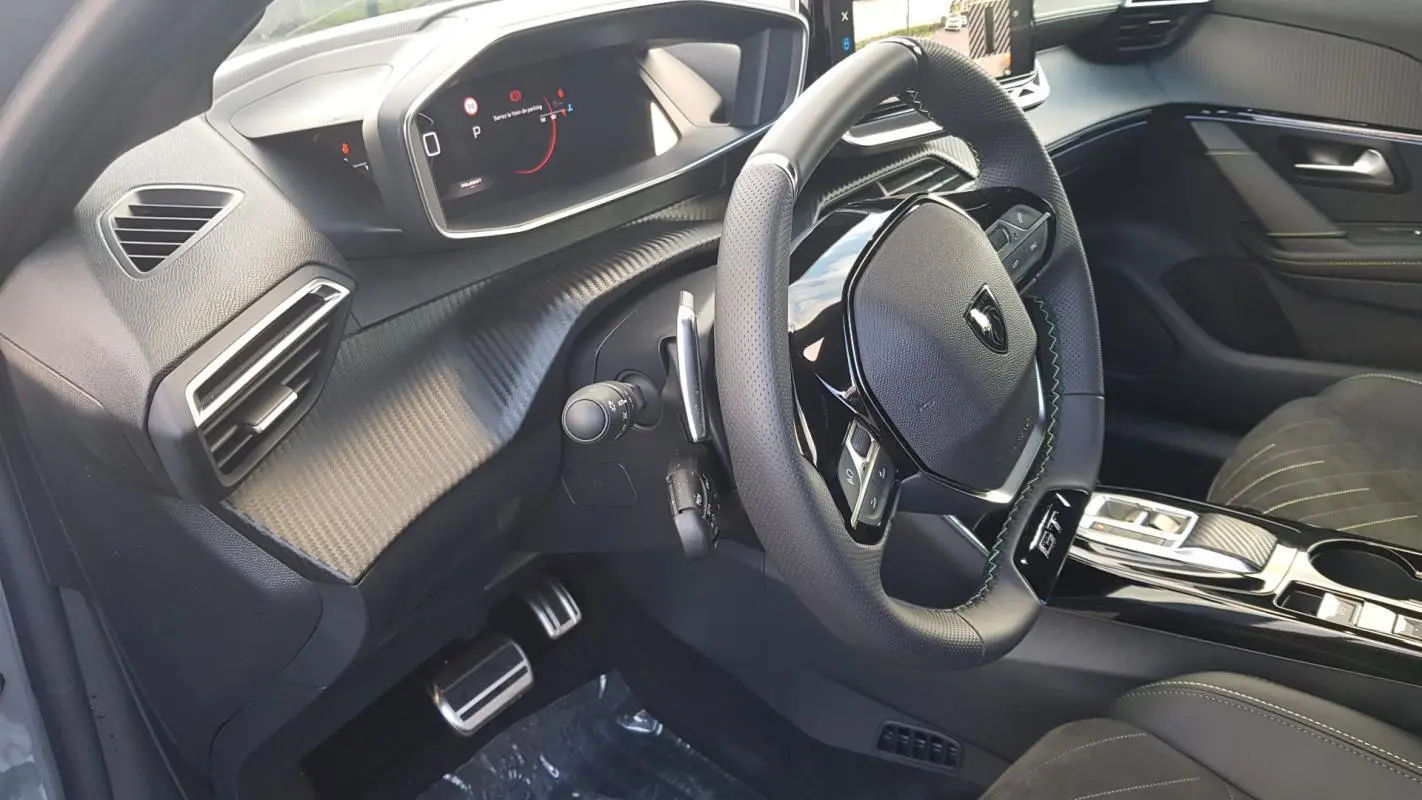 Vue intérieure côté conducteur du Peugeot 208 Hybrid 110 gris, avec volant GT, tableau de bord numérique et finitions carbone.