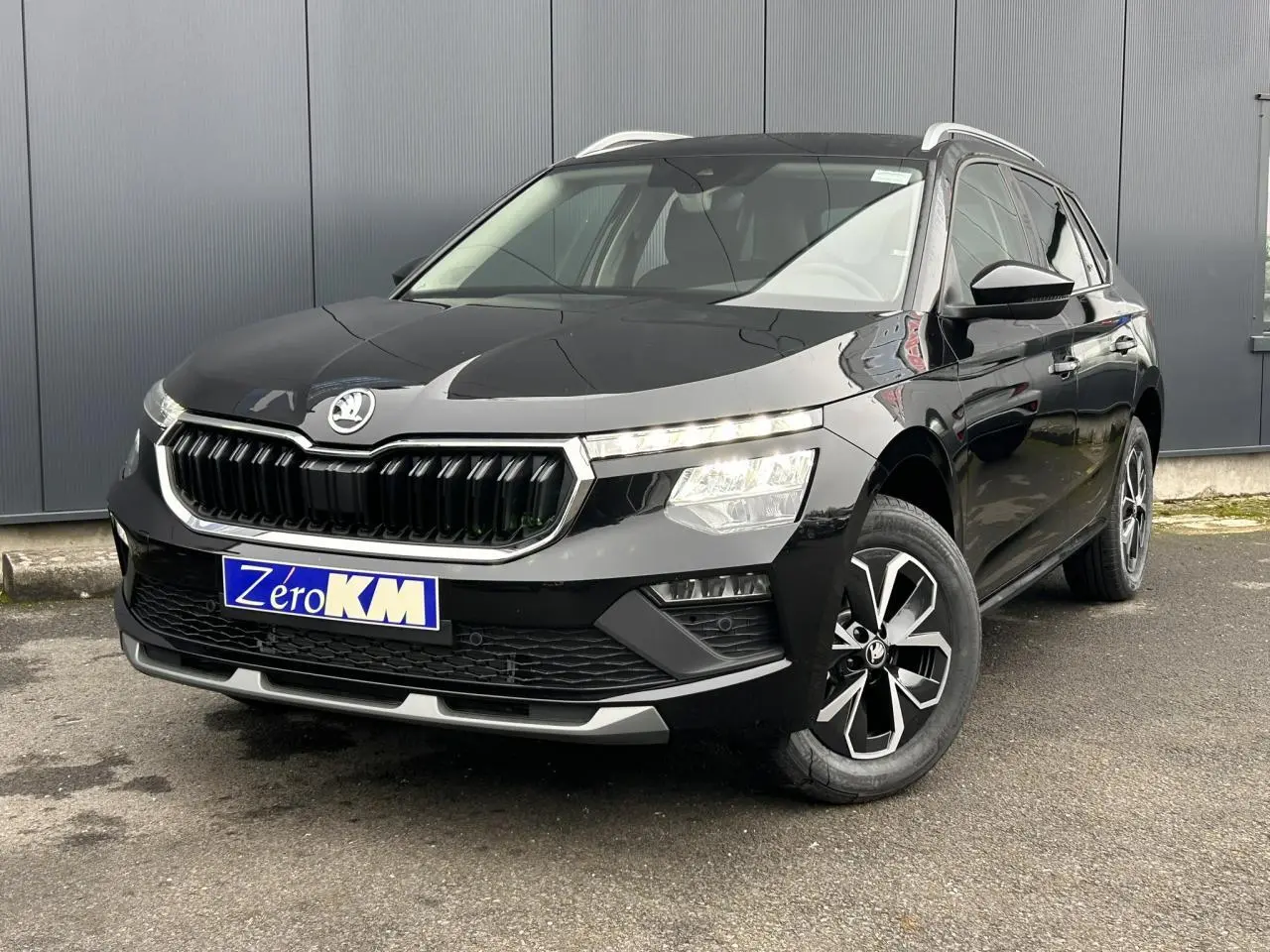 Vue 3/4 avant d'un Skoda Kamiq noir Magic avec jantes alliage et calandre chromée, stationné devant un mur gris.