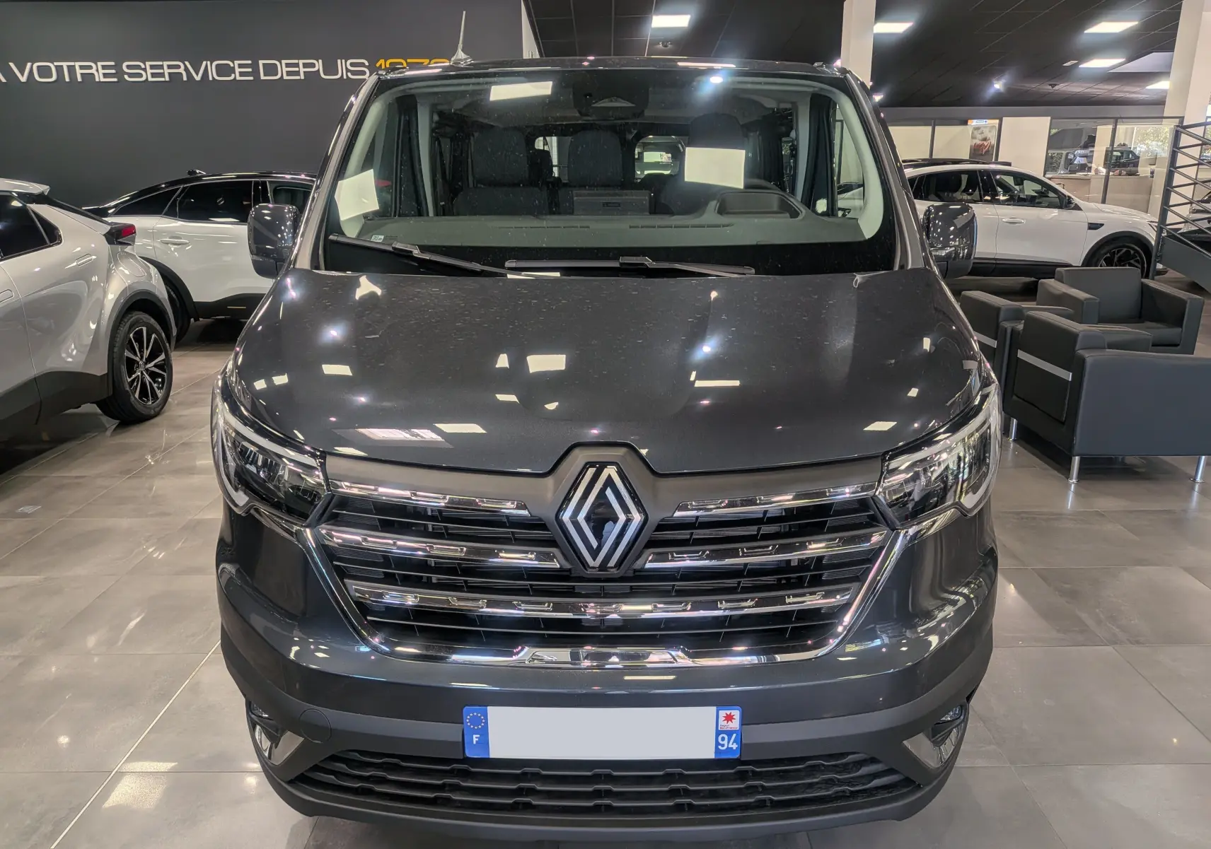 Vue frontale d'un Renault Trafic Combi gris Comète 2025 en showroom, avec calandre chromée et logo Renault bien visible.