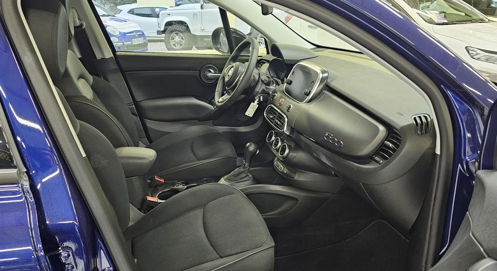 Intérieur du FIAT 500X 2024 bleu, vue côté gauche montrant sièges noirs et tableau de bord moderne avec écran tactile.