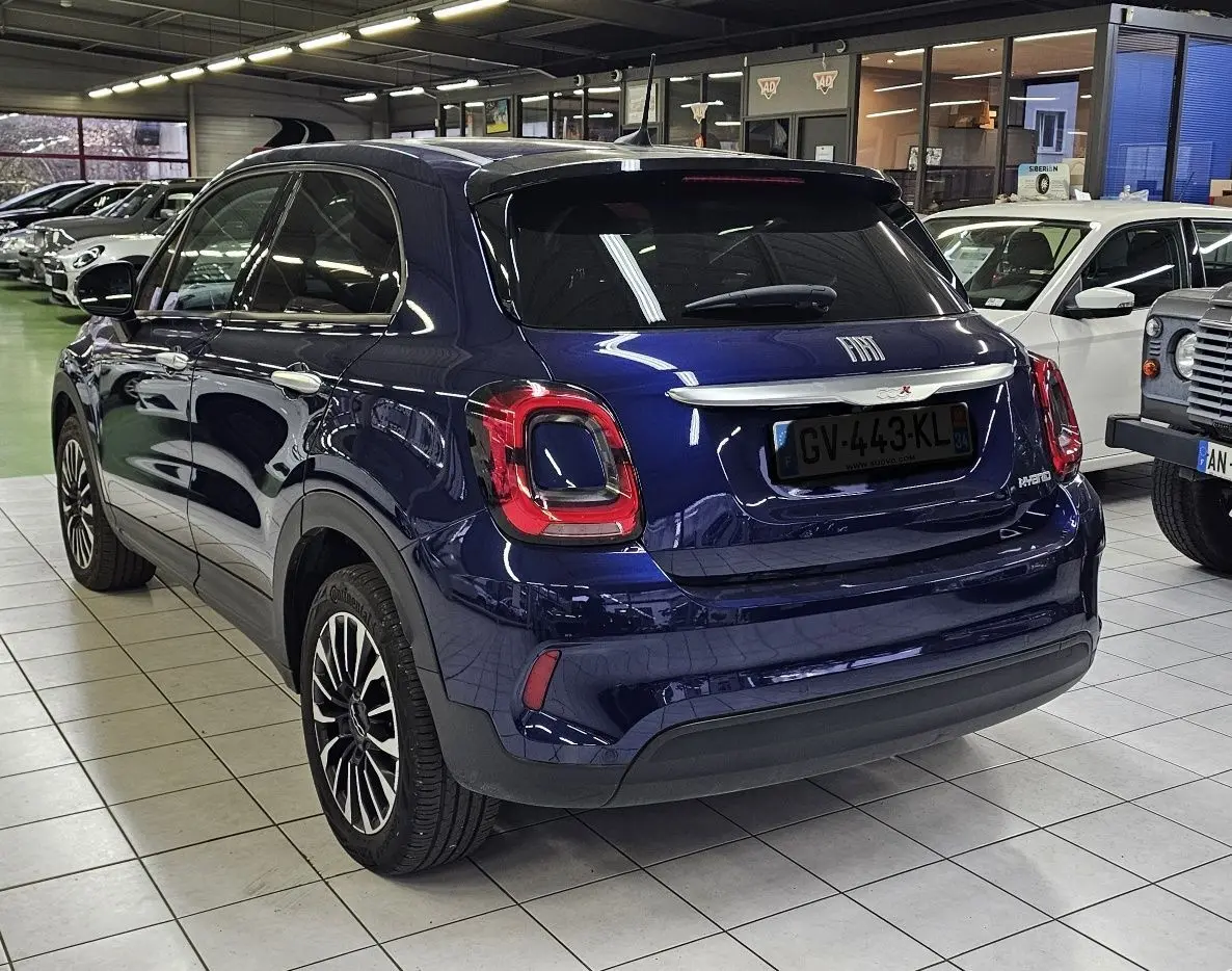 Vue 3/4 arrière d'un FIAT 500X bleu 2024 hybride essence en showroom, avec feux arrière distinctifs et jantes stylées.