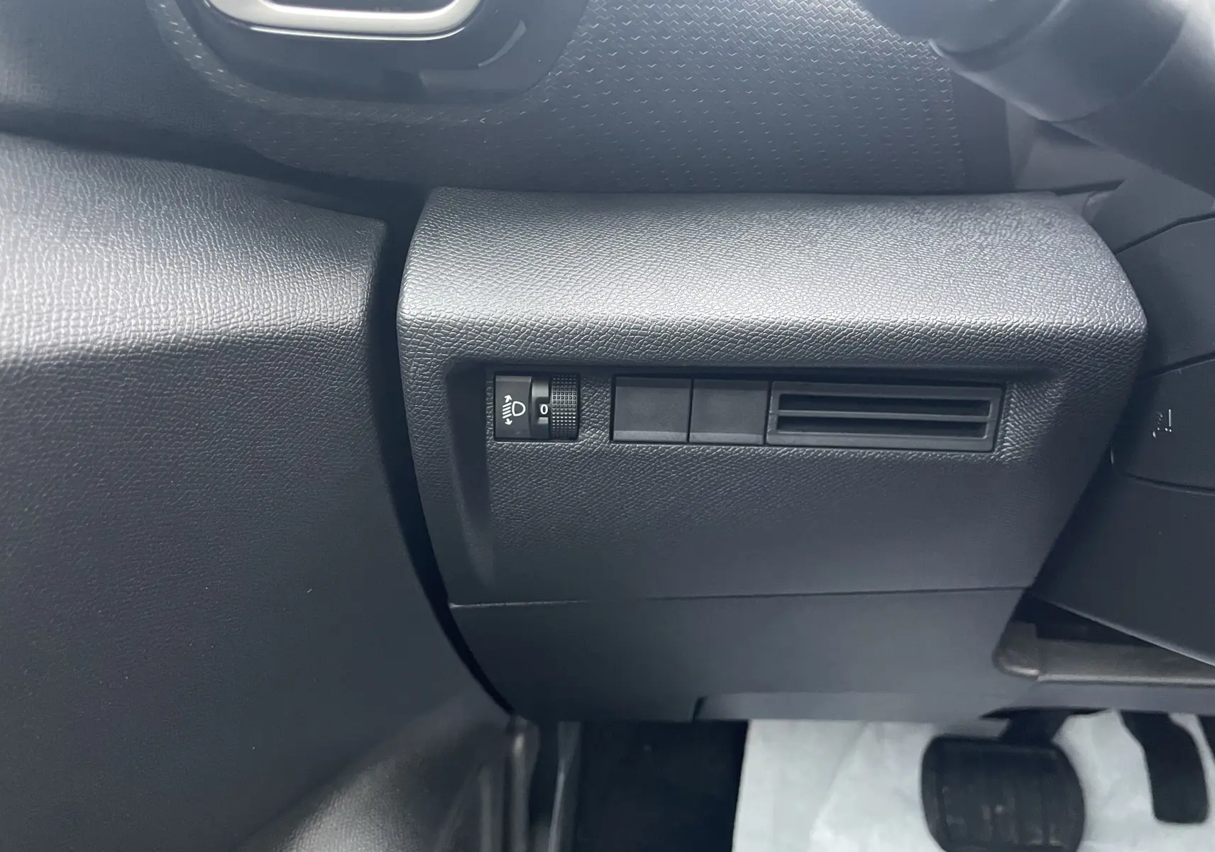 Détail intérieur côté gauche du tableau de bord noir avec commande de réglage des phares sur Citroën C4 hybride 2025.