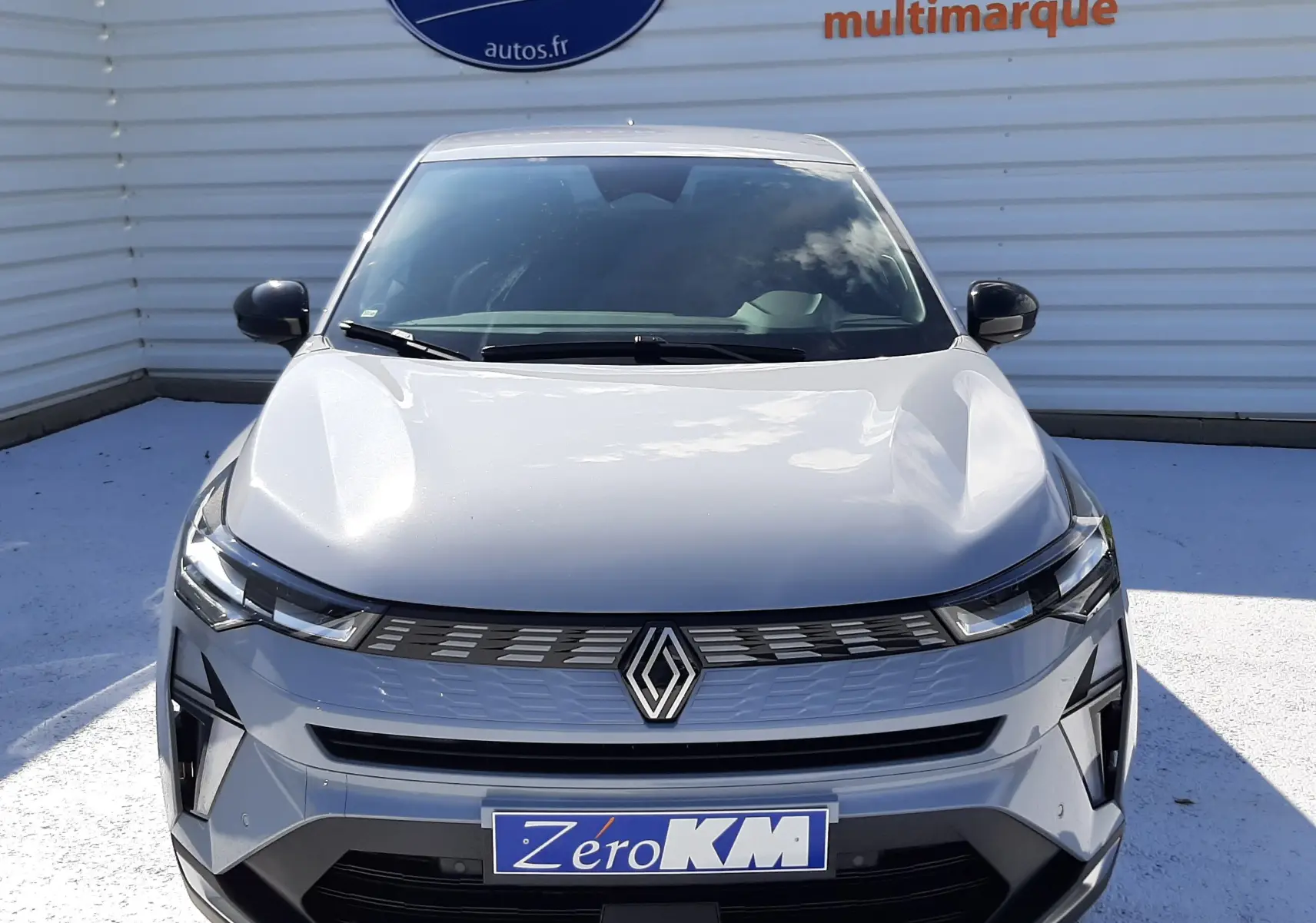 Vue frontale d'une Renault SYMBIOZ gris rafale 2024 avec calandre moderne et logo centré, stationnée devant un mur publicitaire.
