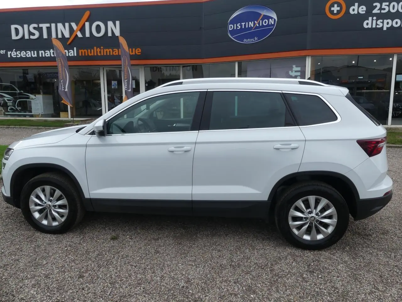 Profil côté gauche d'un SUV SKODA KAROQ blanc 2024 avec jantes alliage 17 pouces devant un concessionnaire Distinxion.