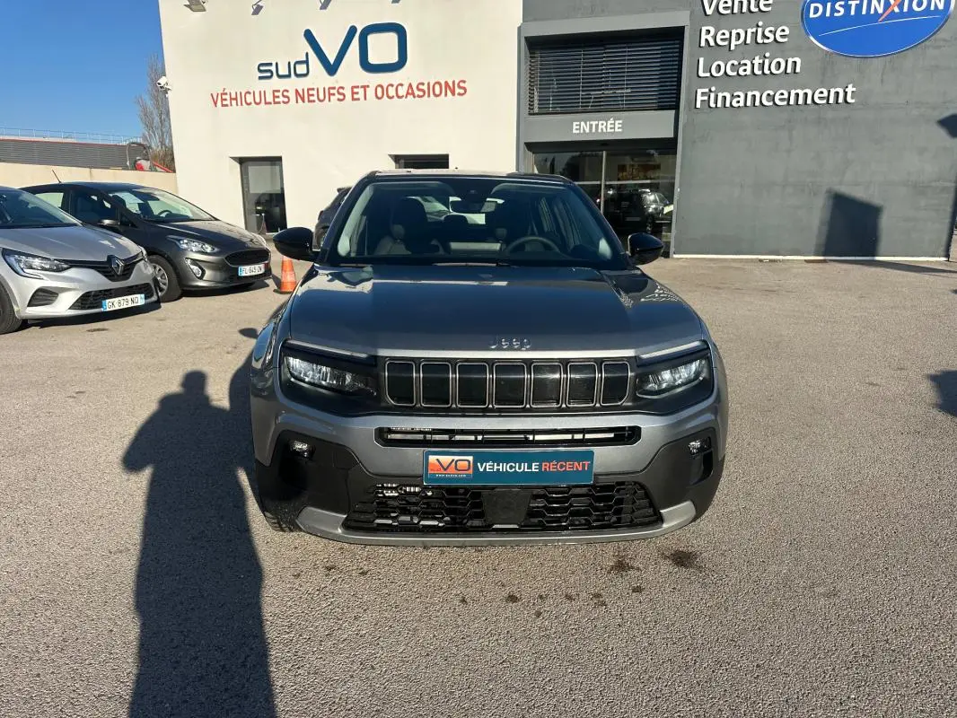 Vue de face d’un Jeep Avenger gris foncé 2025 avec calandre distinctive et phares LED allumés, stationné devant un concessionnaire.
