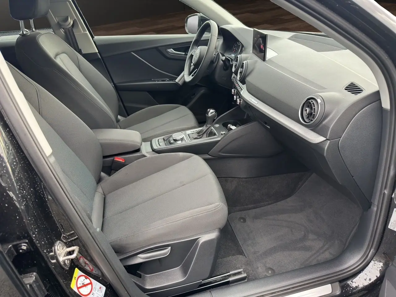 Intérieur côté passager d'un Audi Q2 noir Mito 2023, sièges tissu gris et tableau de bord moderne avec écran GPS.