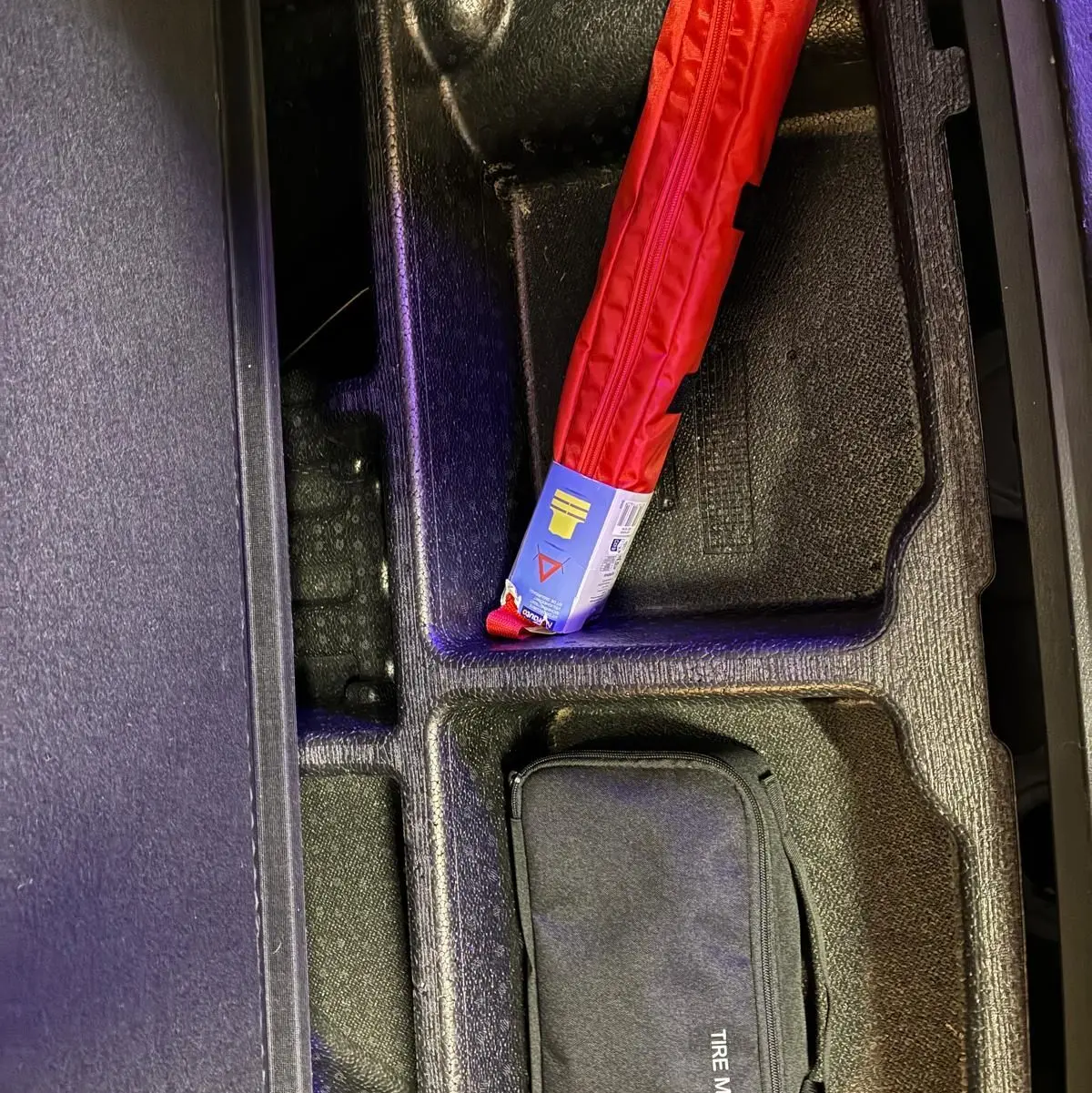 Rangement sous coffre du KIA Sportage hybride 2024 montrant un kit mobilité pneus noir et un triangle de signalisation rouge.