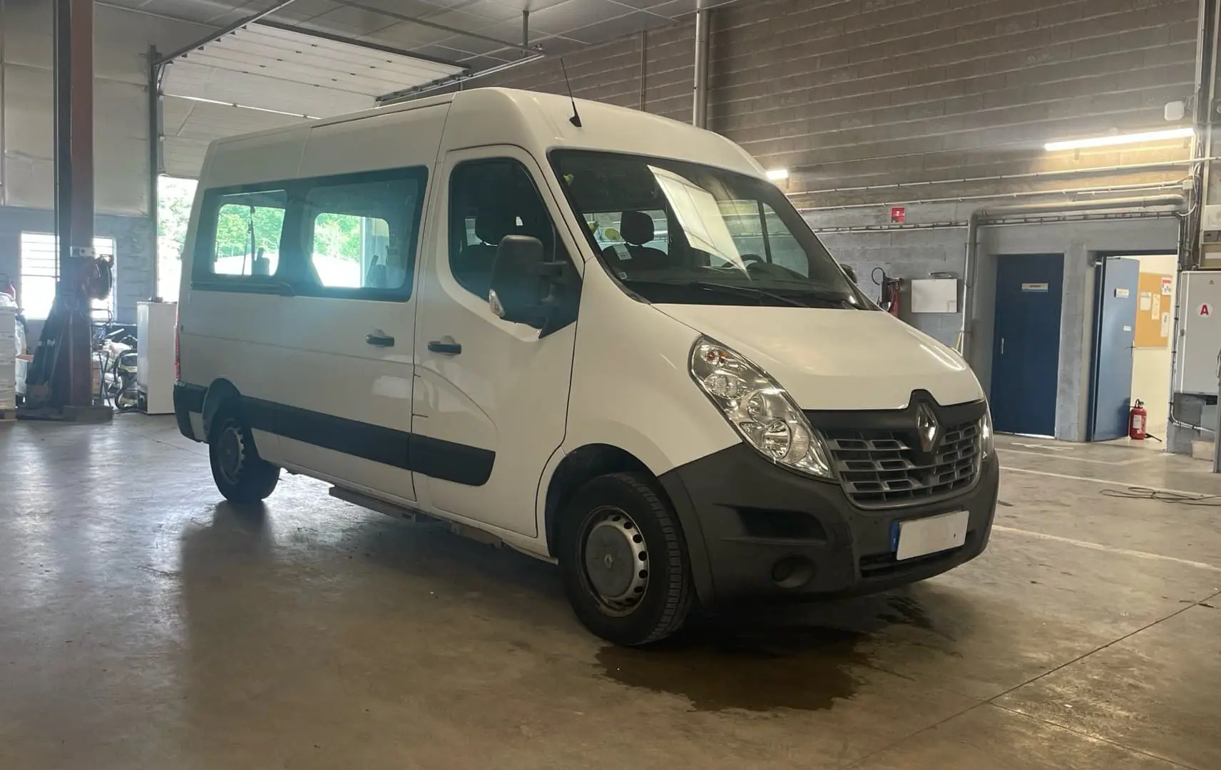 Renault Master blanc en 3/4 avant droit dans un garage, avec grandes fenêtres latérales et marchepied visible.
