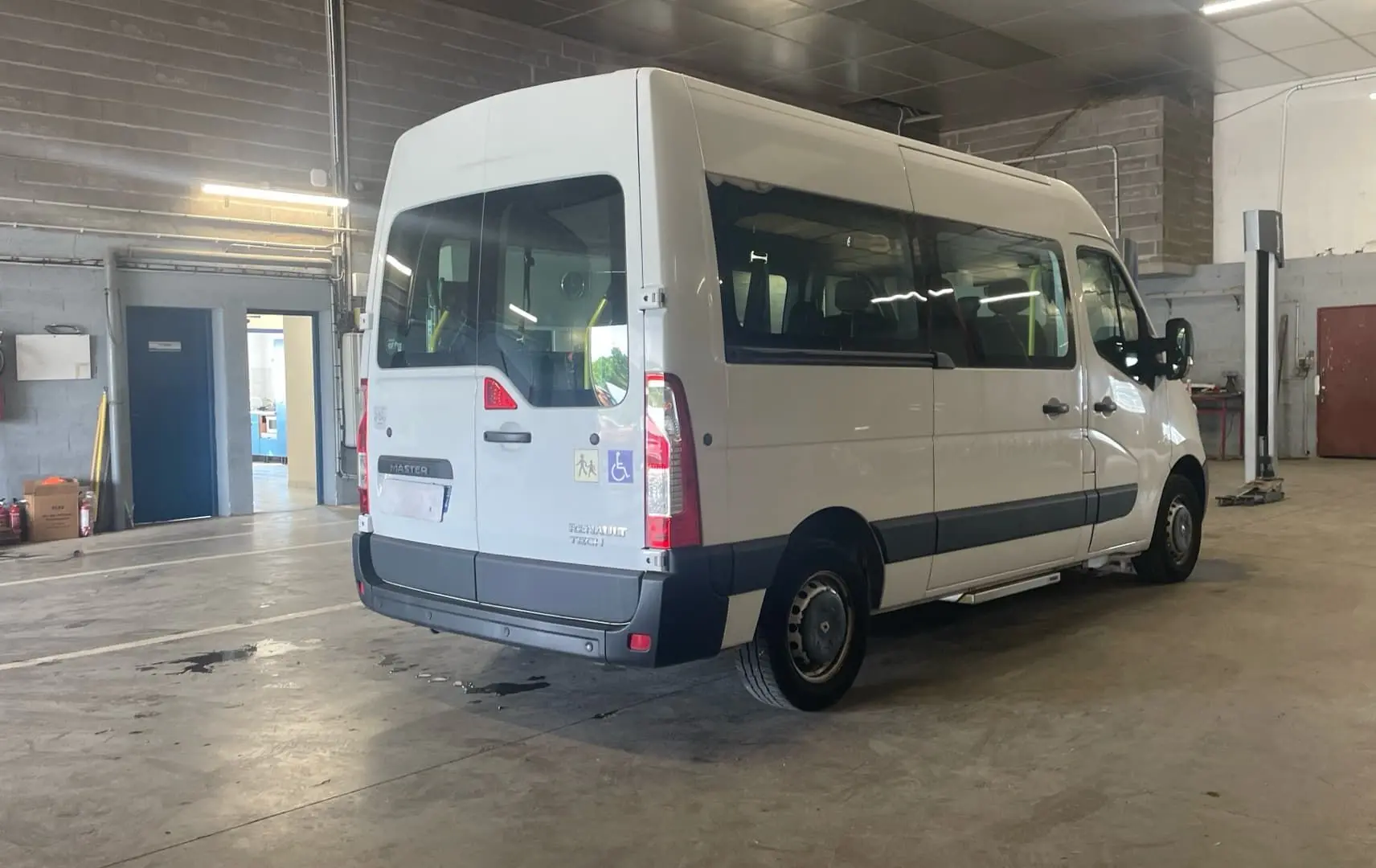 Renault Master blanc en 3/4 arrière droit dans un garage, avec rampe arrière et logo handicap visible sur la porte.