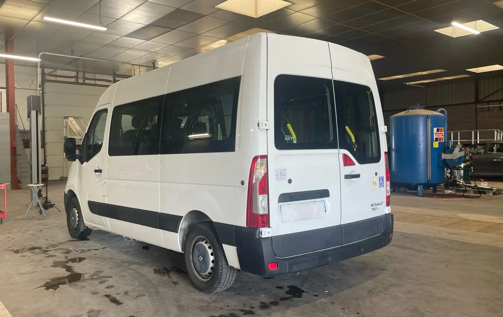 Renault Master blanc vu en 3/4 arrière droit dans un atelier, avec rampe arrière et vitres latérales teintées.