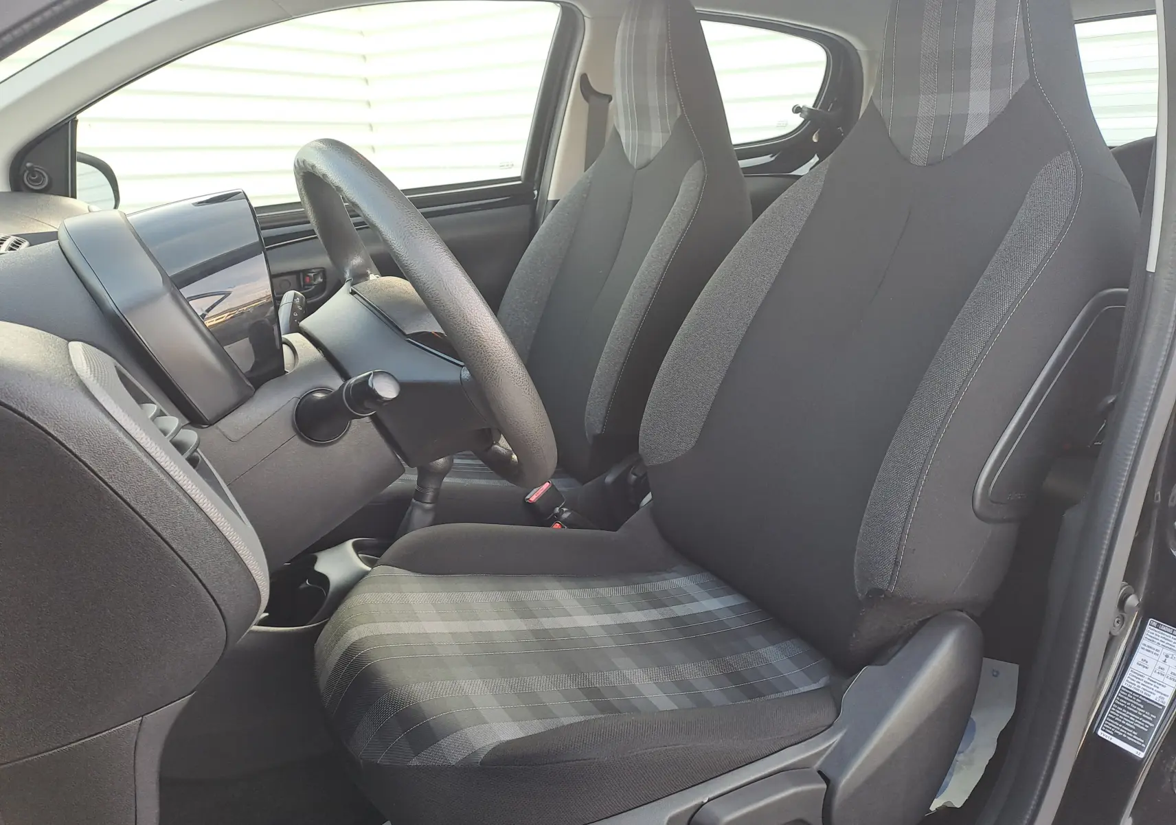 Intérieur avant droit de la Peugeot 108 noire 2020, sièges tissu noir et gris à motifs carreaux, volant et tableau de bord visibles.