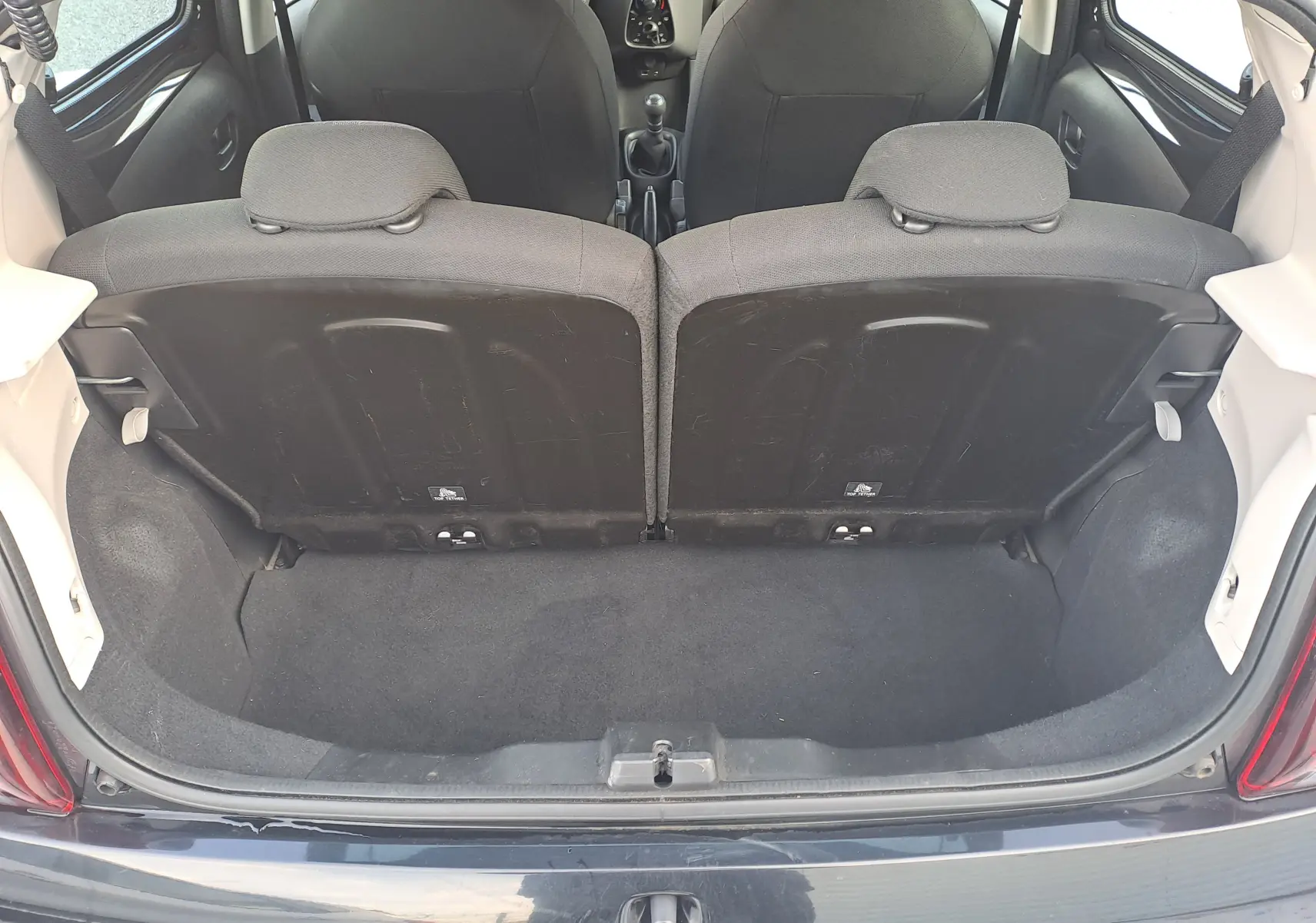 Coffre ouvert vu de l'arrière d'une Peugeot 108 noire, montrant les dossiers arrière et l'espace de chargement.