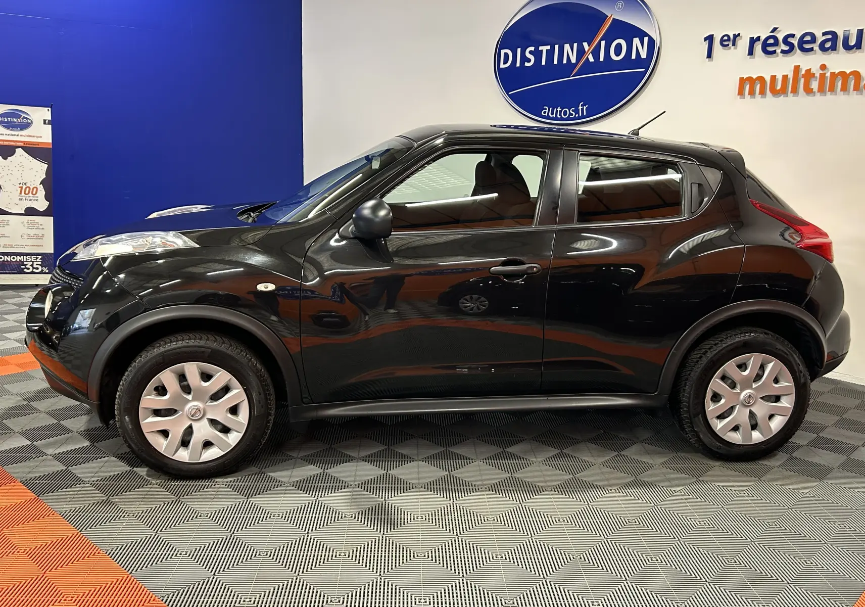 Profil côté gauche d'un Nissan Juke noir métal de 2012 avec attelage visible, posé en intérieur sur sol damier.