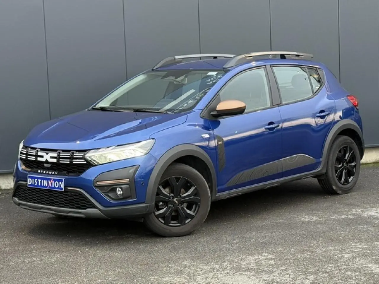 Dacia Sandero Stepway Eco-G 100 Extrême bleu Iron en 3/4 avant droit avec barres de toit et jantes noires.