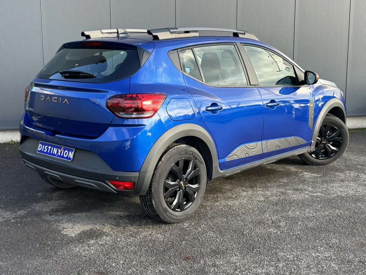 Vue 3/4 arrière droite d'une Dacia Sandero Stepway Bleu Iron avec jantes noires et barres de toit grises.