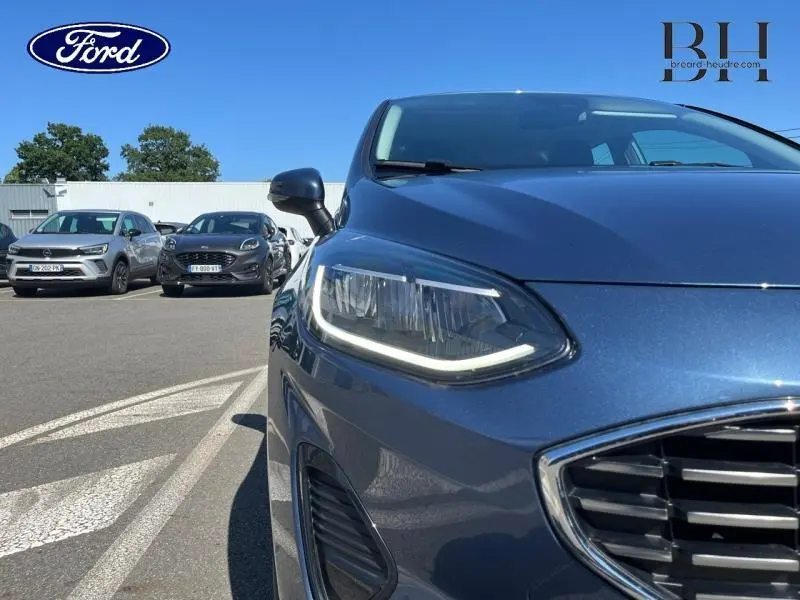 Gros plan sur l'avant droit d'une Ford Fiesta bleu azur métallisée, mettant en valeur son phare LED et la calandre chromée.